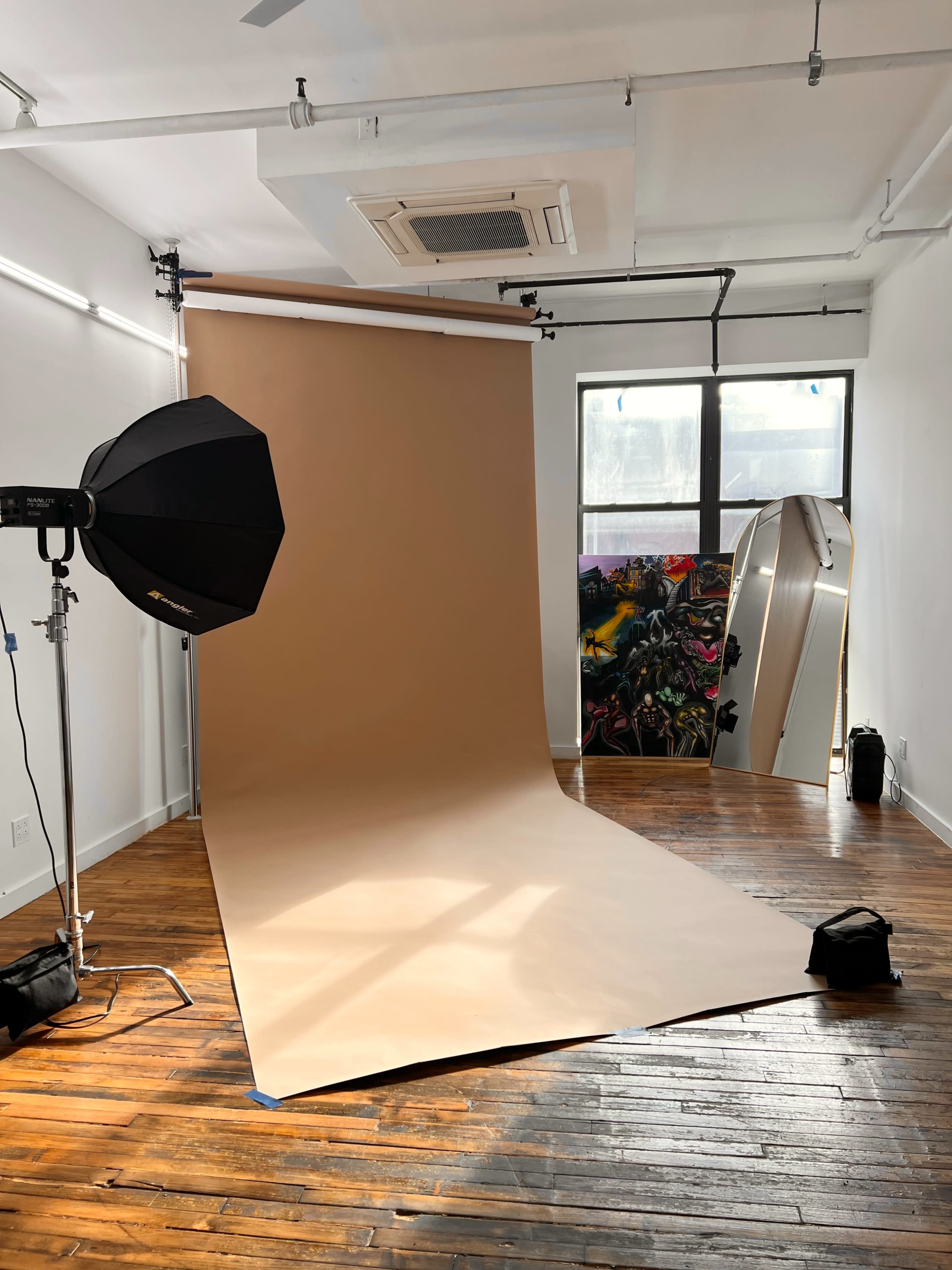 The image shows a photography studio with a large beige backdrop, a softbox light, and a mirror, along with colorful wall art visible through a window.
