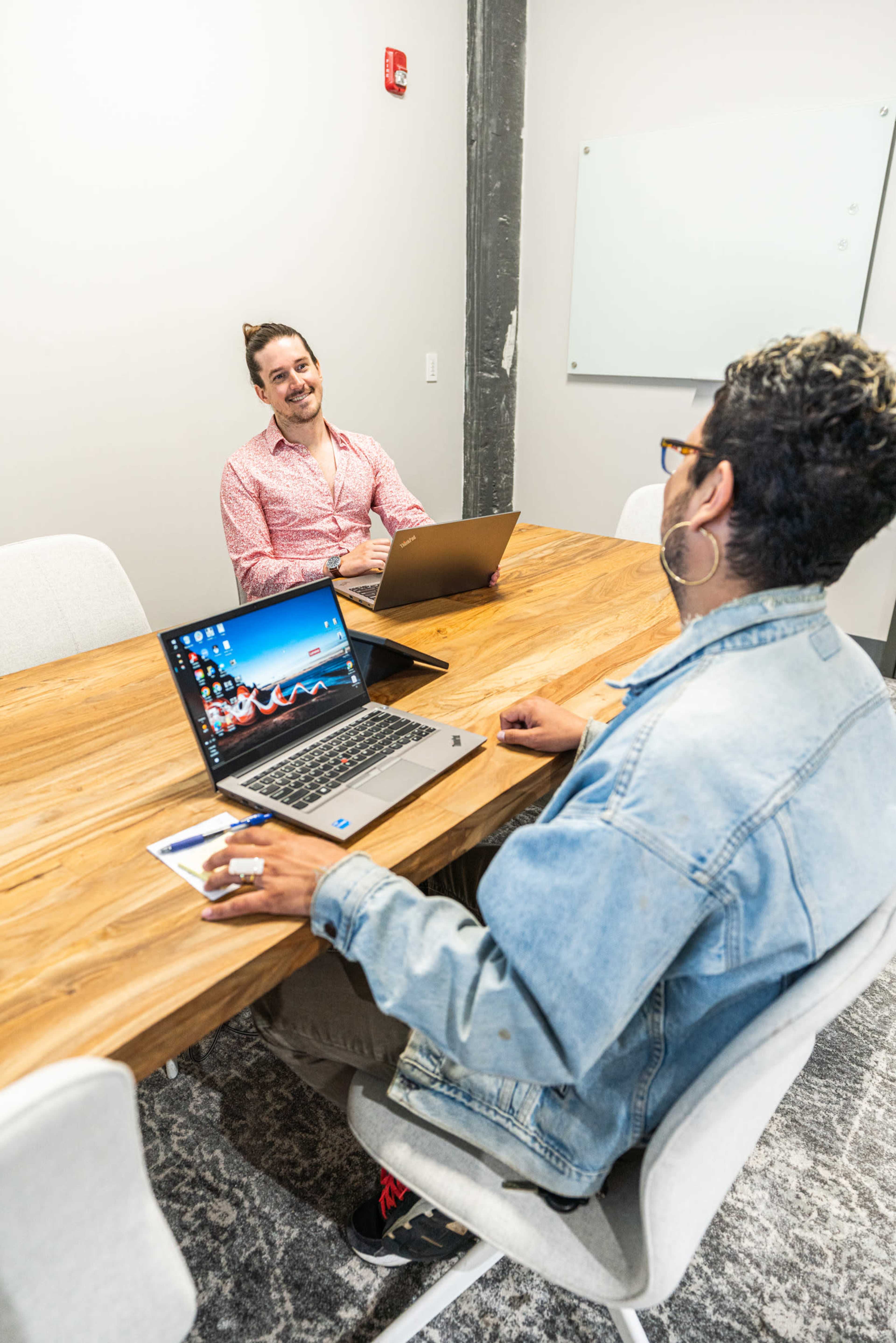 Two people are sitting at a wooden table in a meeting room, each using a laptop and engaging in conversation.