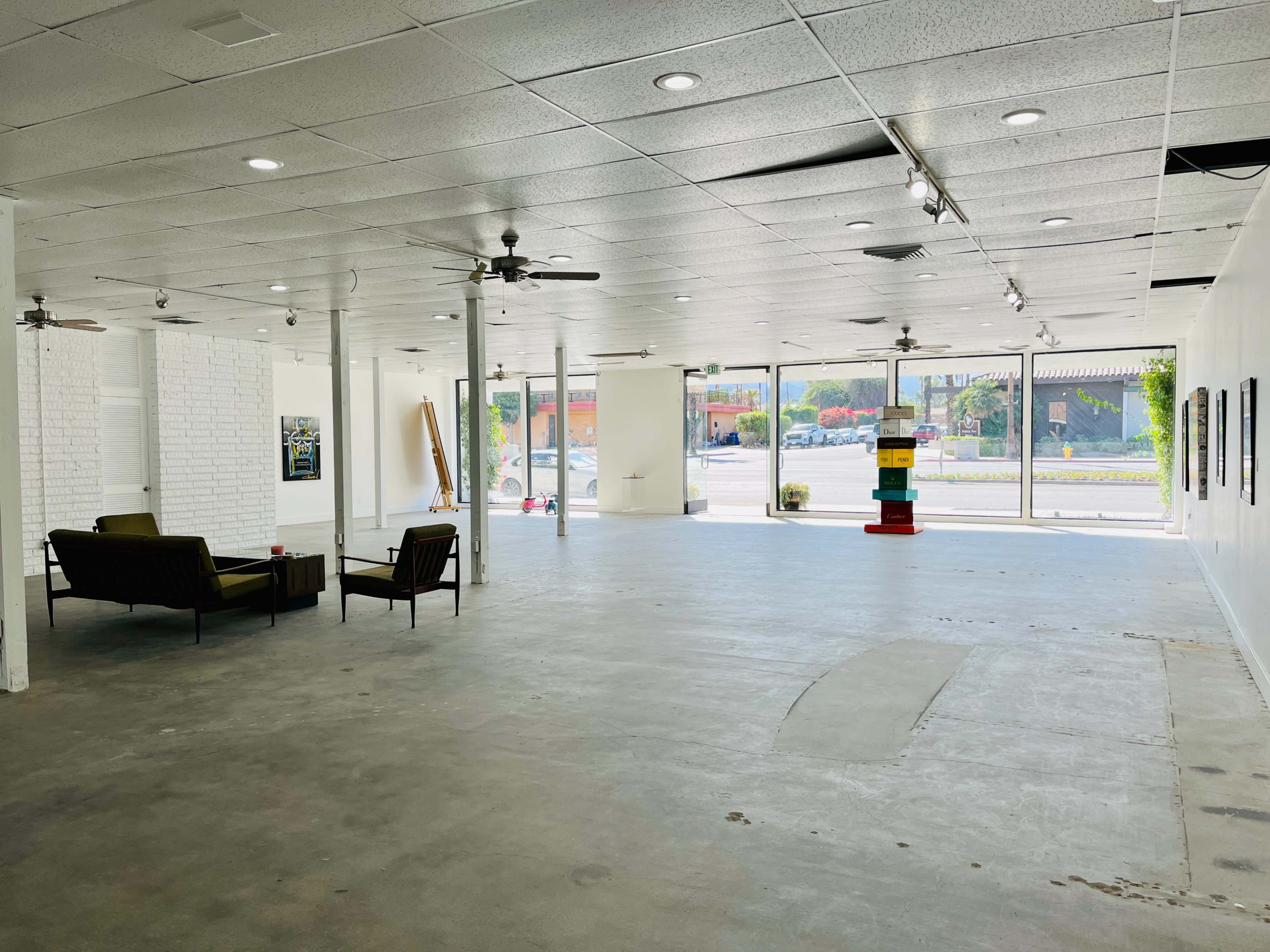 A spacious, empty gallery with concrete floors, large front windows, and minimal seating along one side.