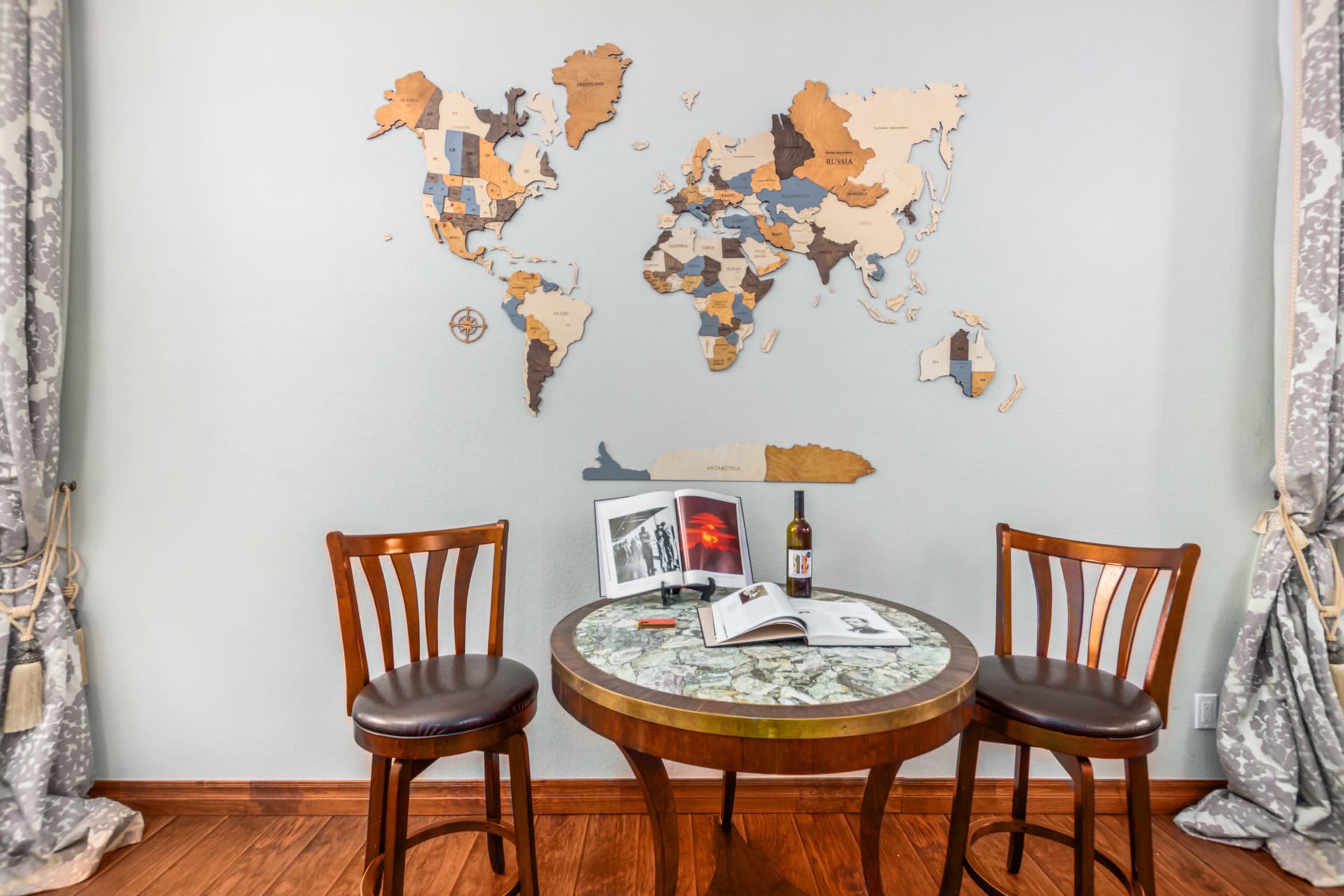 The scene features a round table with two wooden chairs, a bottle, and books, set against a wall decorated with a wooden world map.