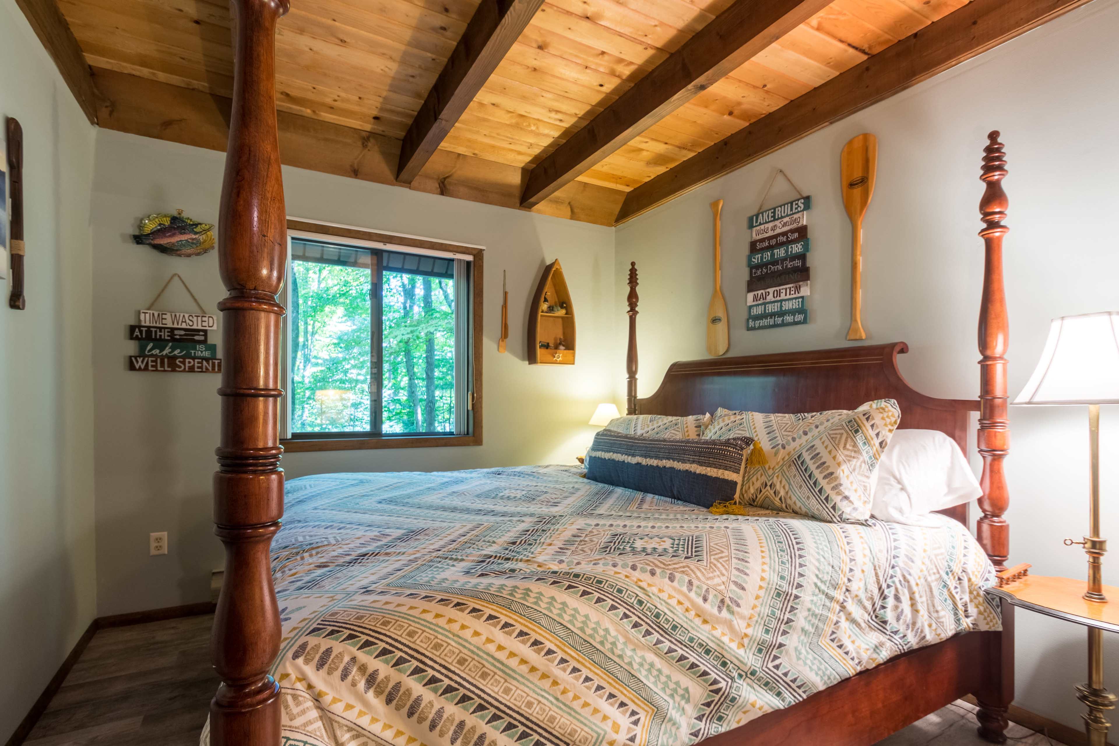 A cozy bedroom features a wooden bed with a patterned comforter, wooden beams on the ceiling, and a window overlooking a wooded area.