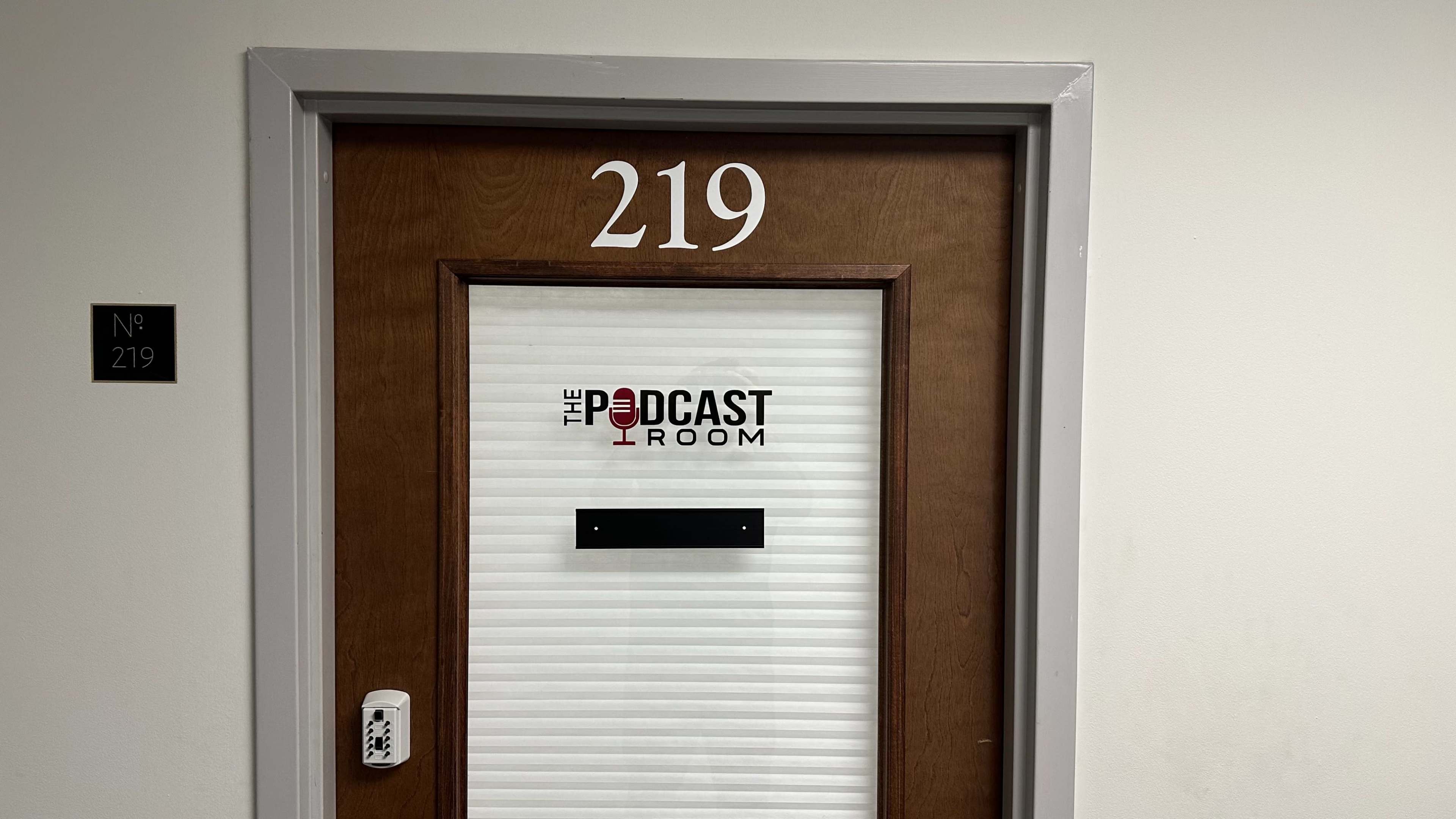 The image shows a door labeled "219" with a sign reading "THE PODCAST ROOM" and a red microphone graphic.
