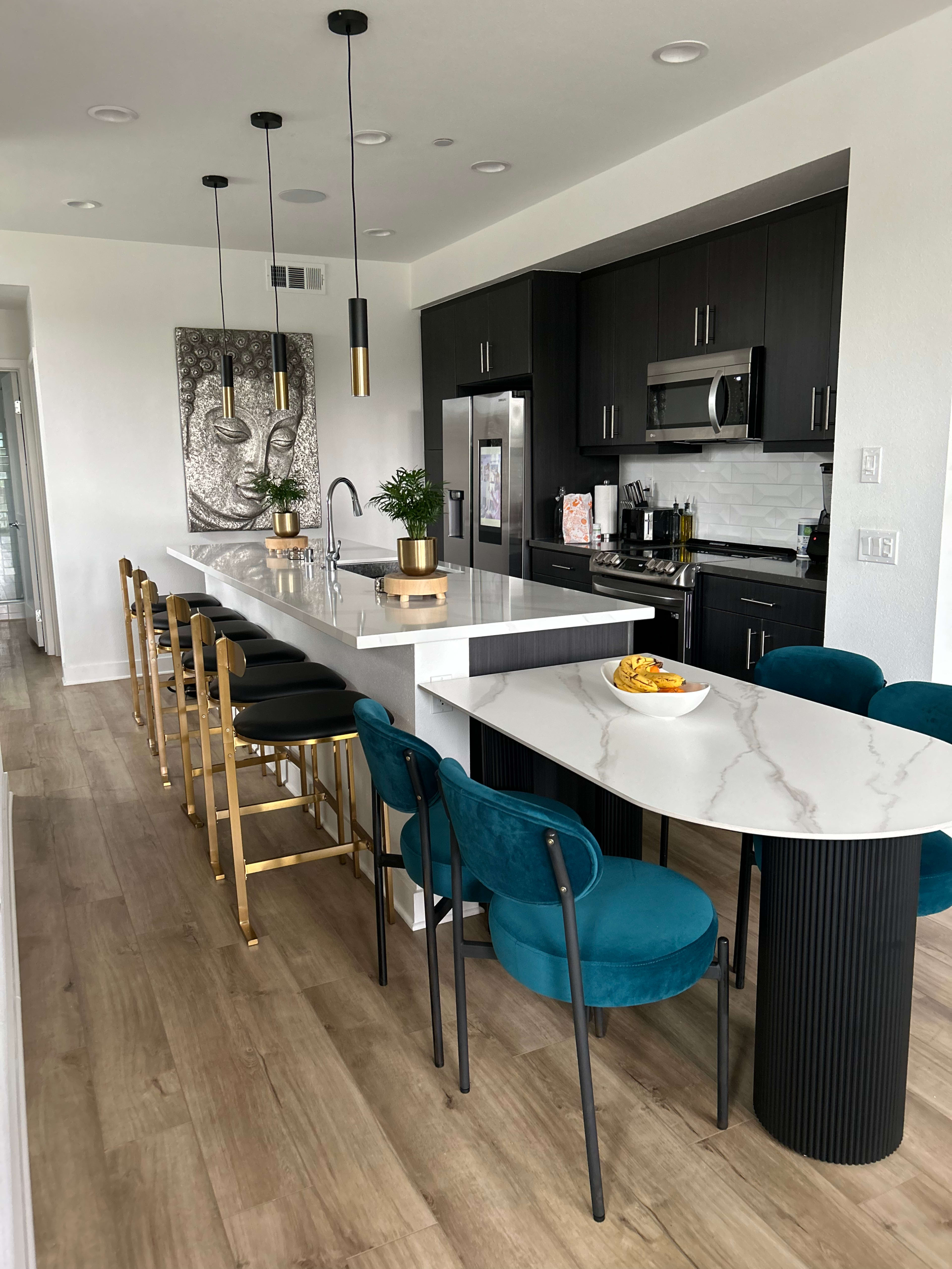 The image shows a modern kitchen with dark cabinets, a large island with white marble countertops, and stylish bar stools, along with a round dining table and teal upholstered chairs.