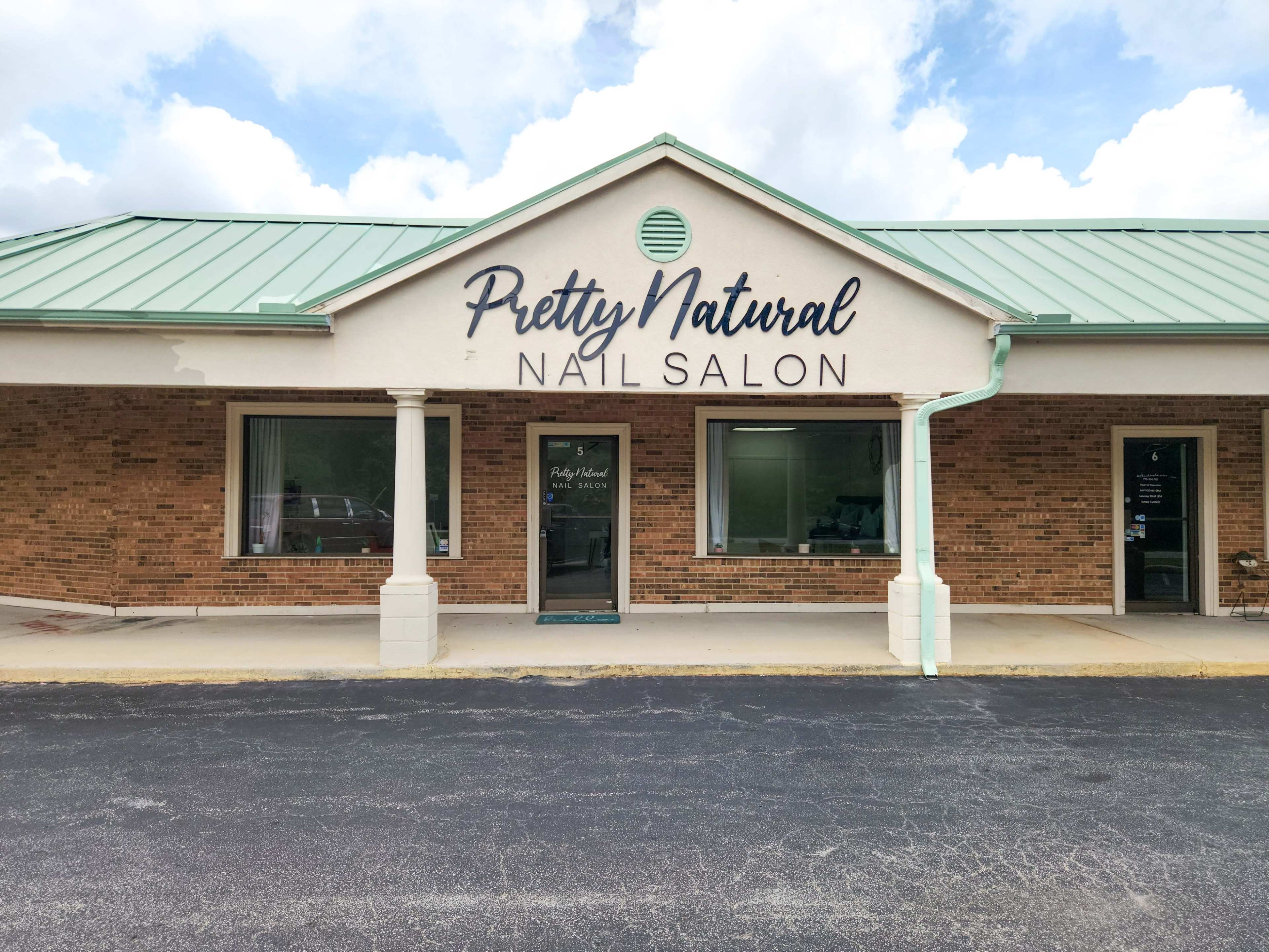 The image shows the exterior façade of a nail salon named "Pretty Natural" with a brick building and a green metal roof.