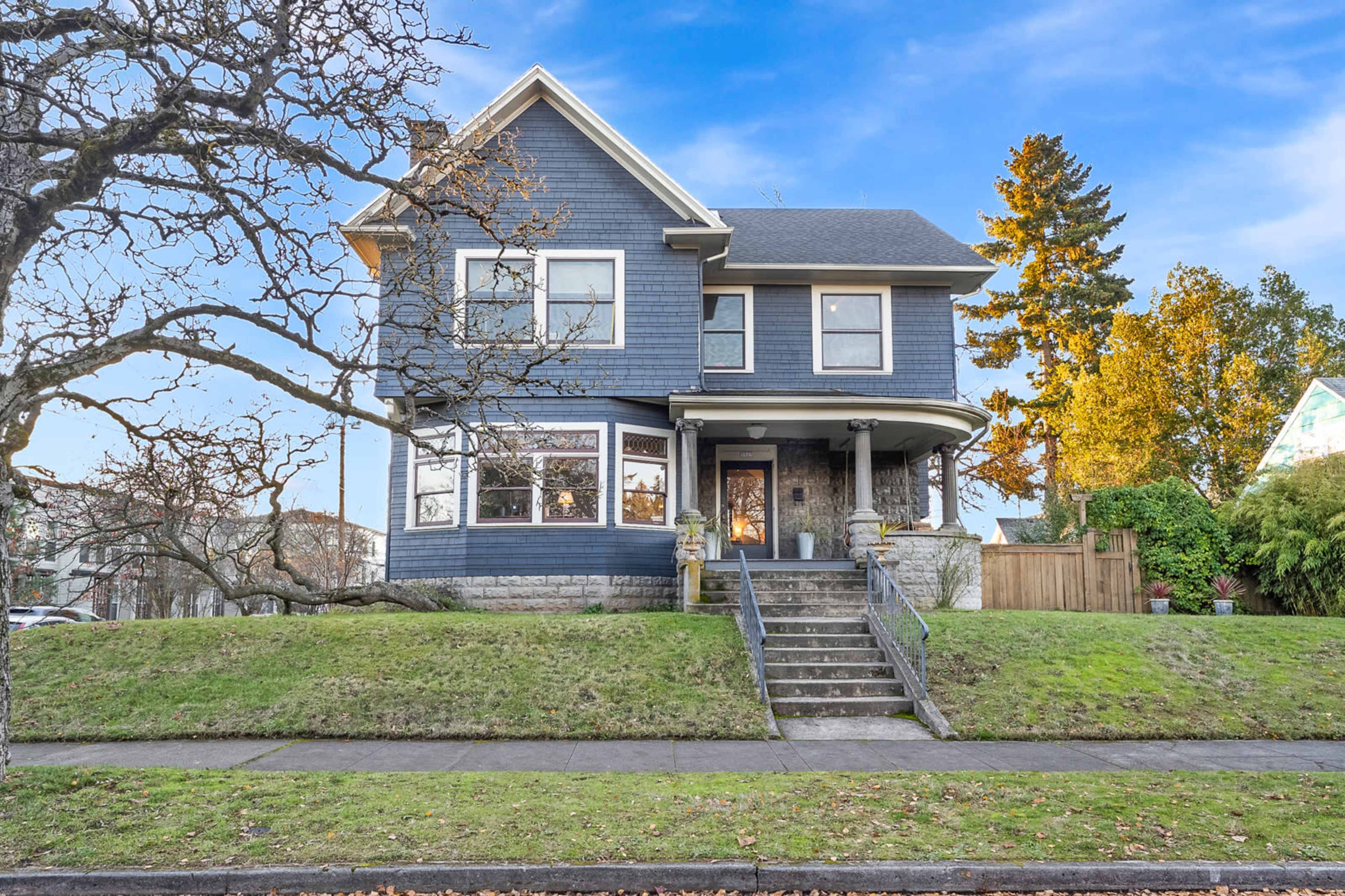 A two-story blue house with a front porch and large windows is situated on a green lawn, bordered by a tree and a sidewalk.