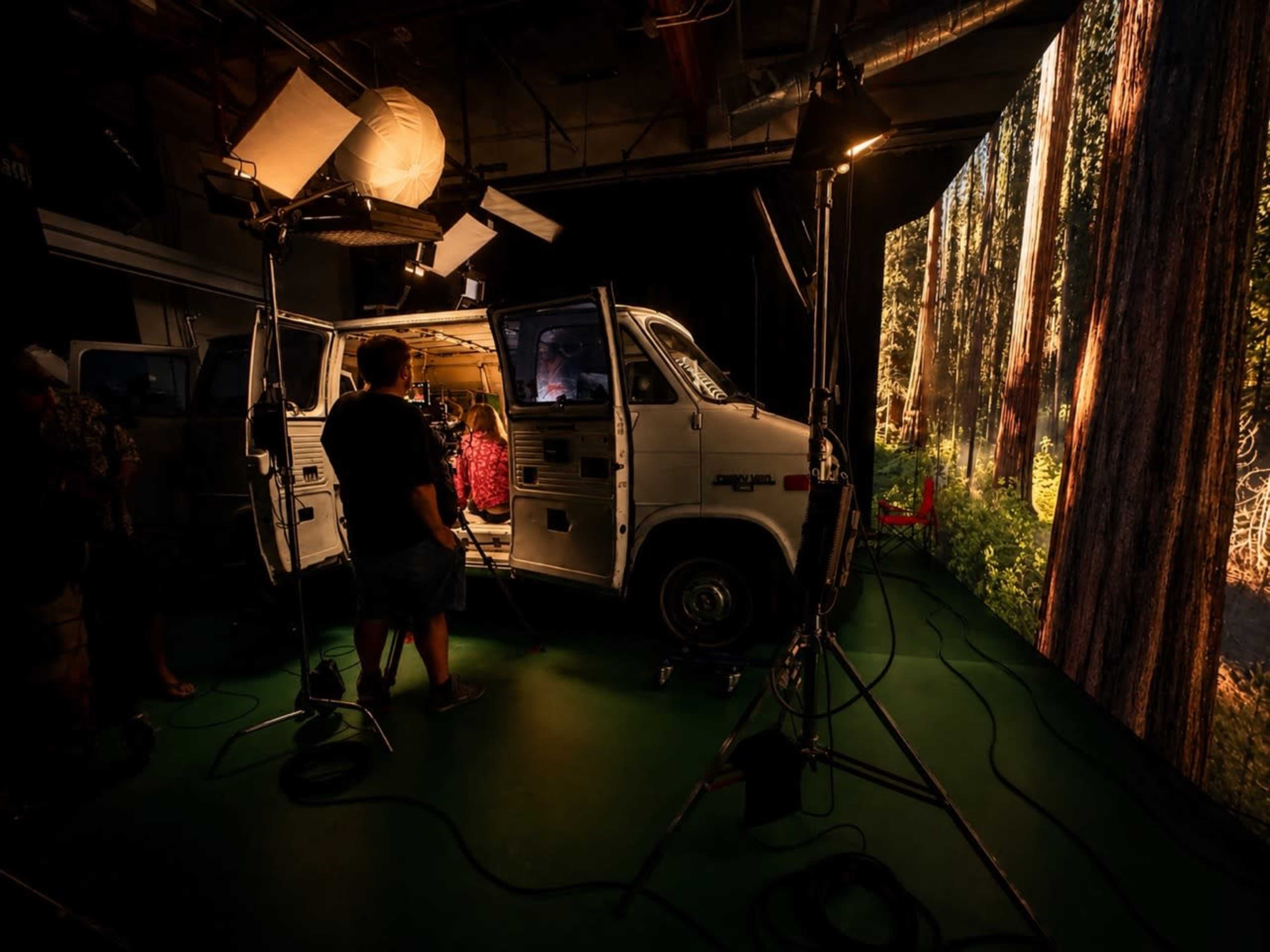 A film crew sets up equipment around a vintage van in a studio, with a backdrop of forest scenery projected on a large screen.