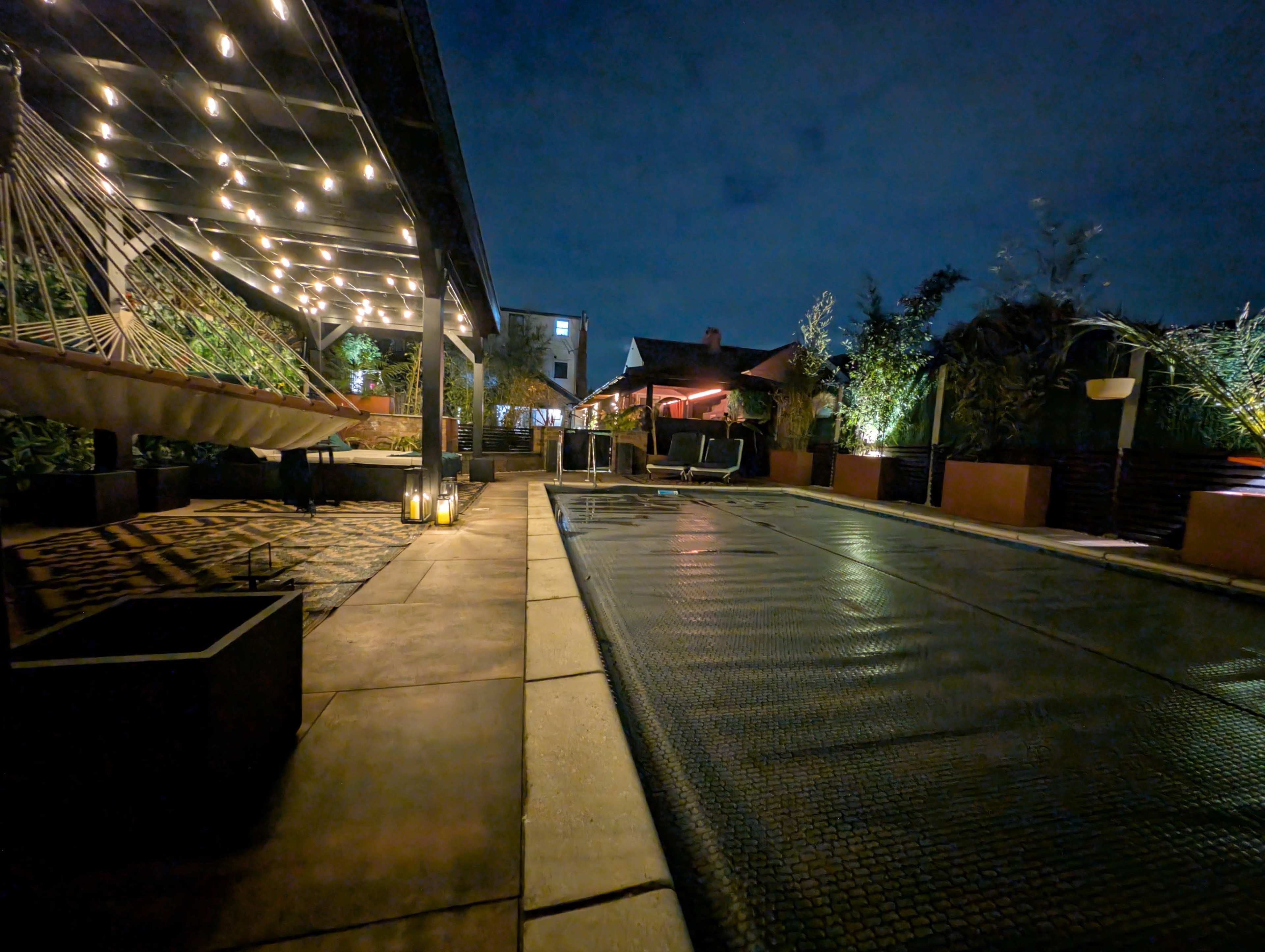 The image shows a dimly lit outdoor area with a pool covered by a net, surrounded by greenery and a seating area with hammocks and decorative lights.