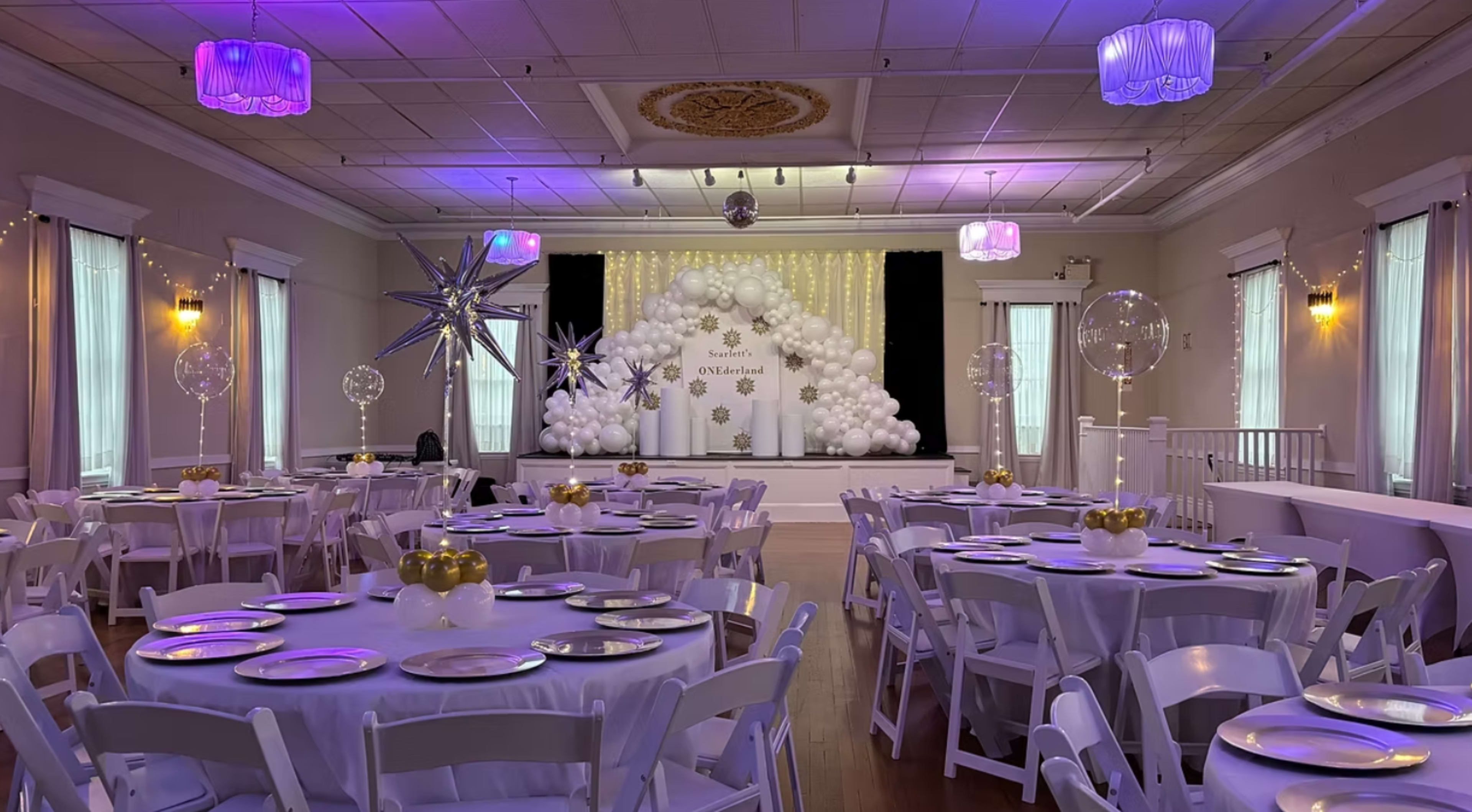 The image depicts a decorated event space with round tables set for dining, a stage adorned with white balloons, and ambient lighting in shades of purple.