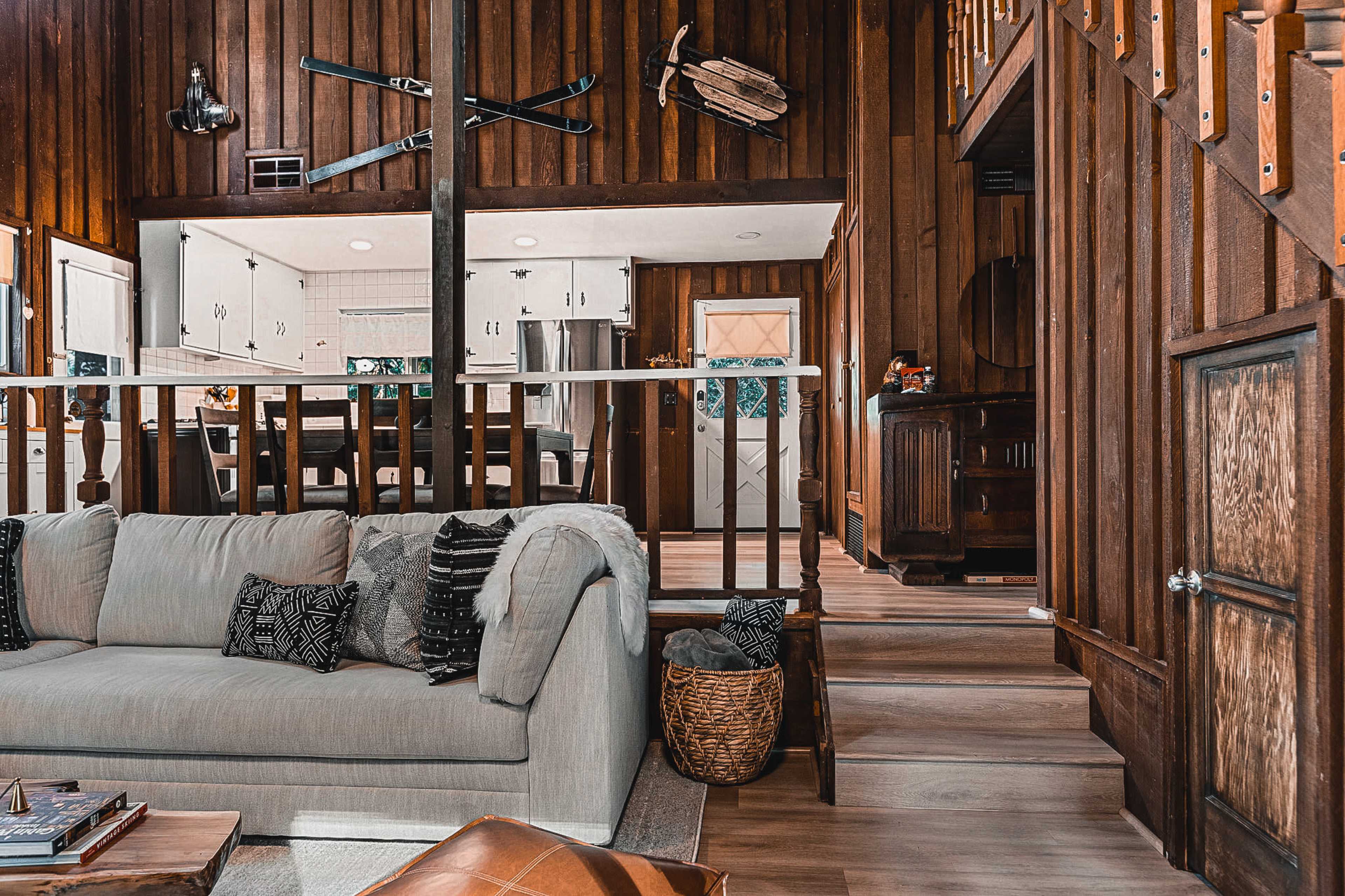 A cozy interior of a wooden cabin with a living area featuring a light-colored sofa, a staircase leading to an upper level, and a kitchen visible in the background.