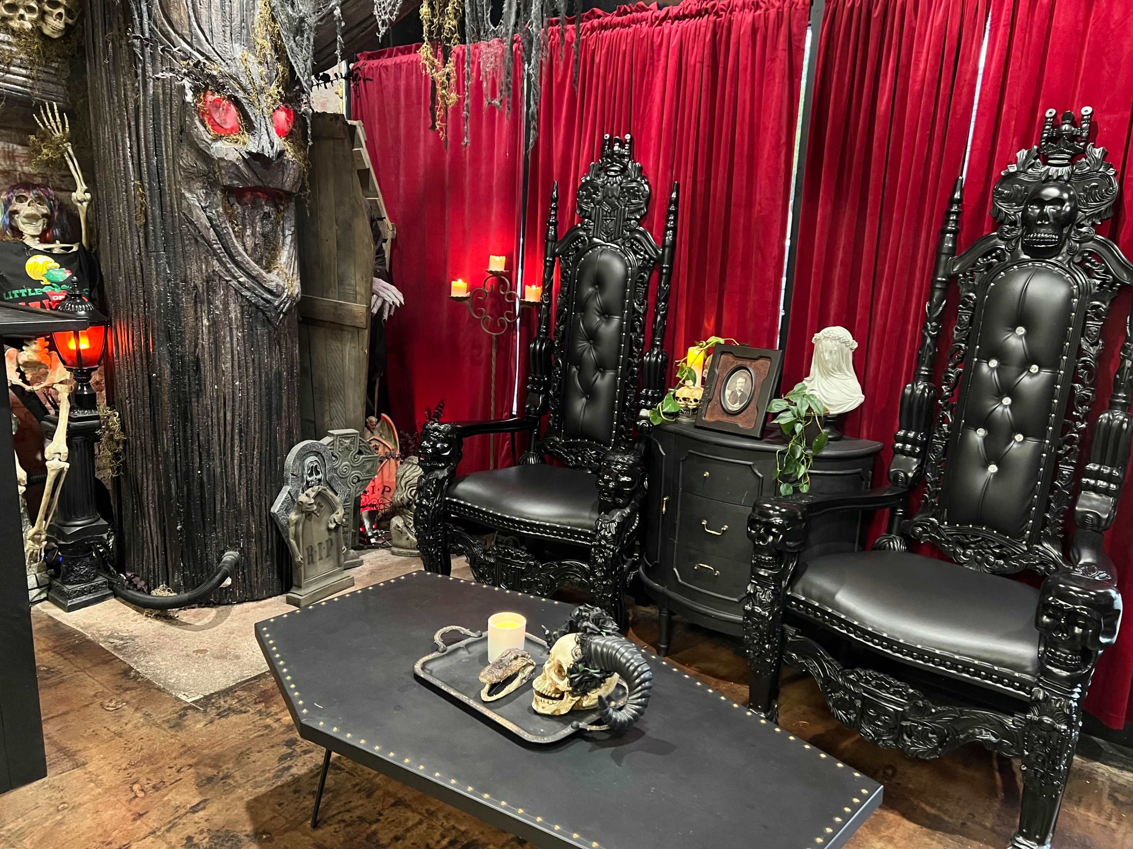 A dark-themed room with two ornate black thrones, a black coffee table, and red velvet curtains, surrounded by skeletal decorations and eerie props.