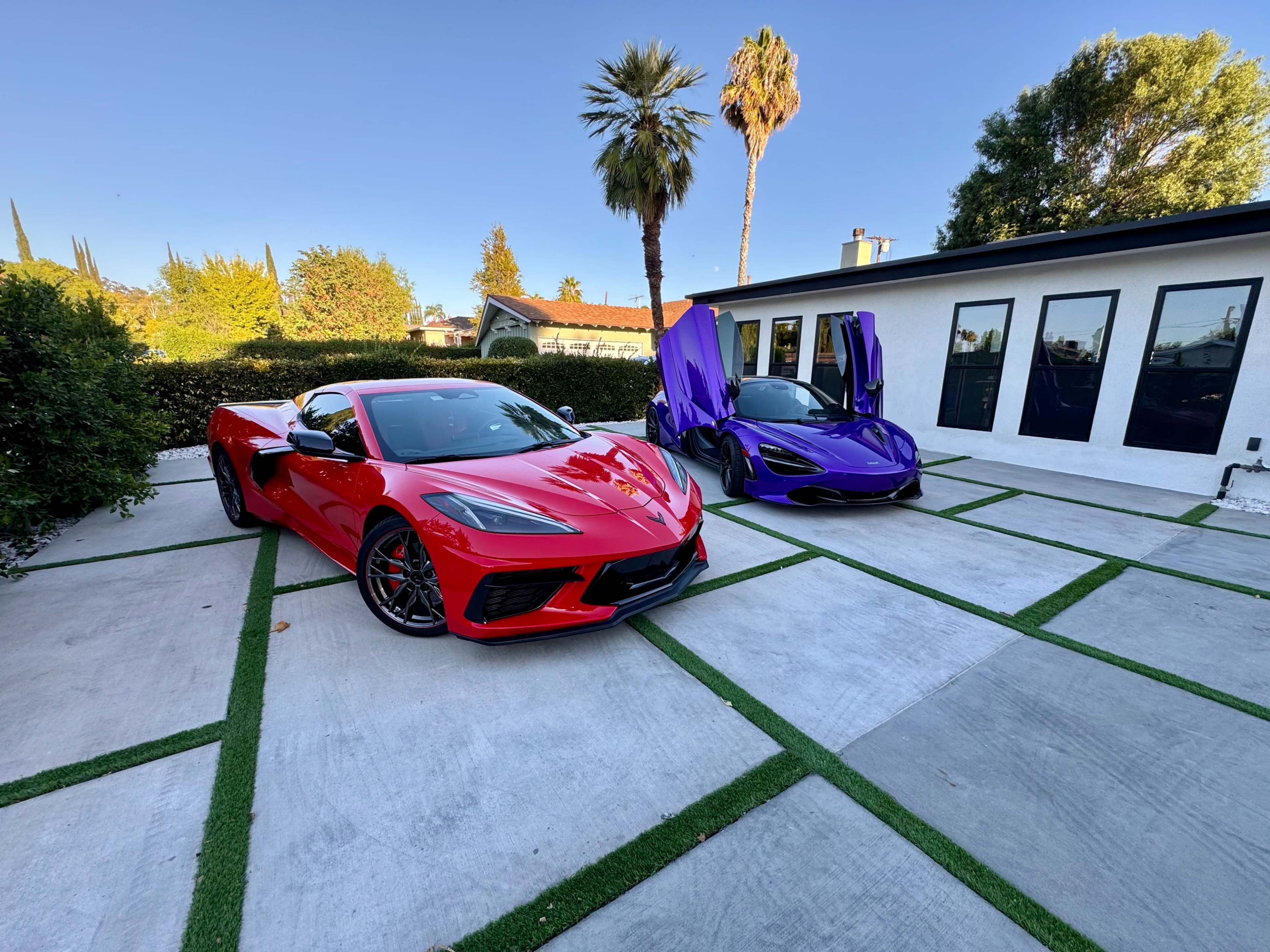 A red C8 Corvette and a purple McLaren are parked on a concrete driveway flanked by palm trees and a modern house.