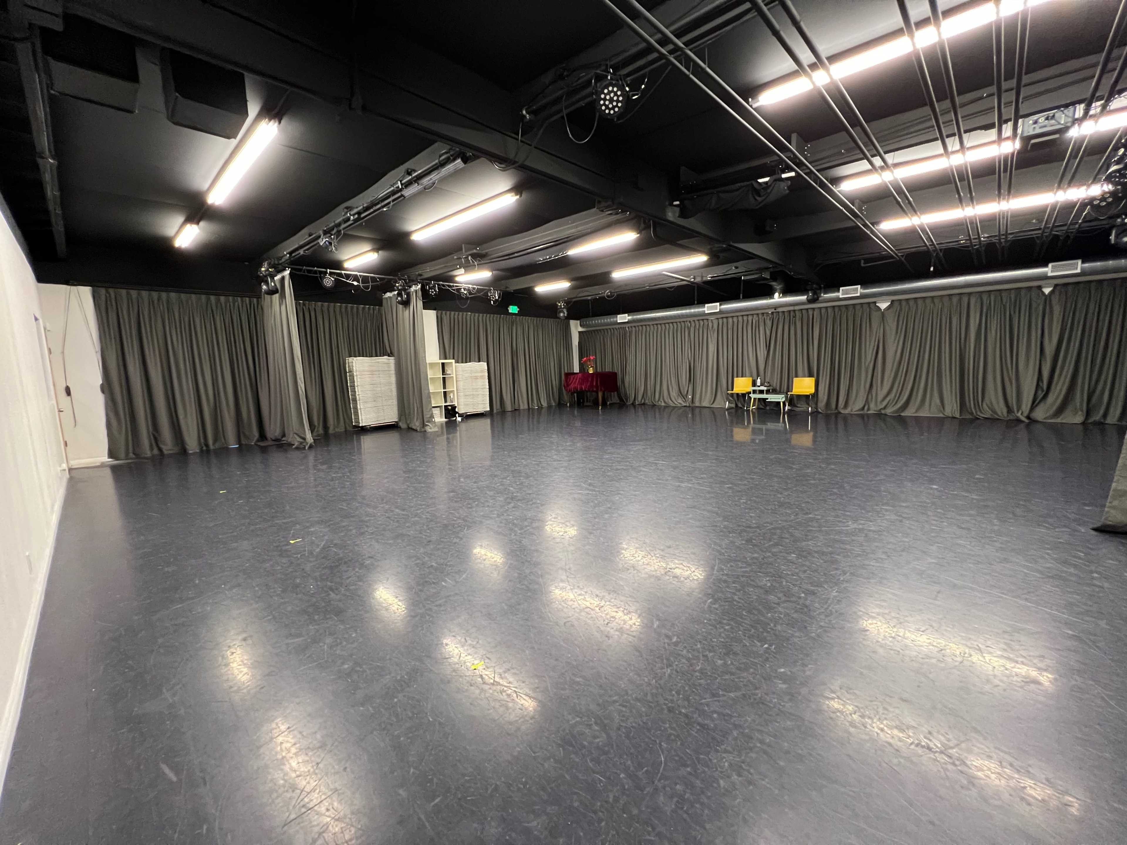 The image shows an empty rehearsal studio with black flooring, white walls, and gray curtains, featuring two yellow chairs and a table at one end.