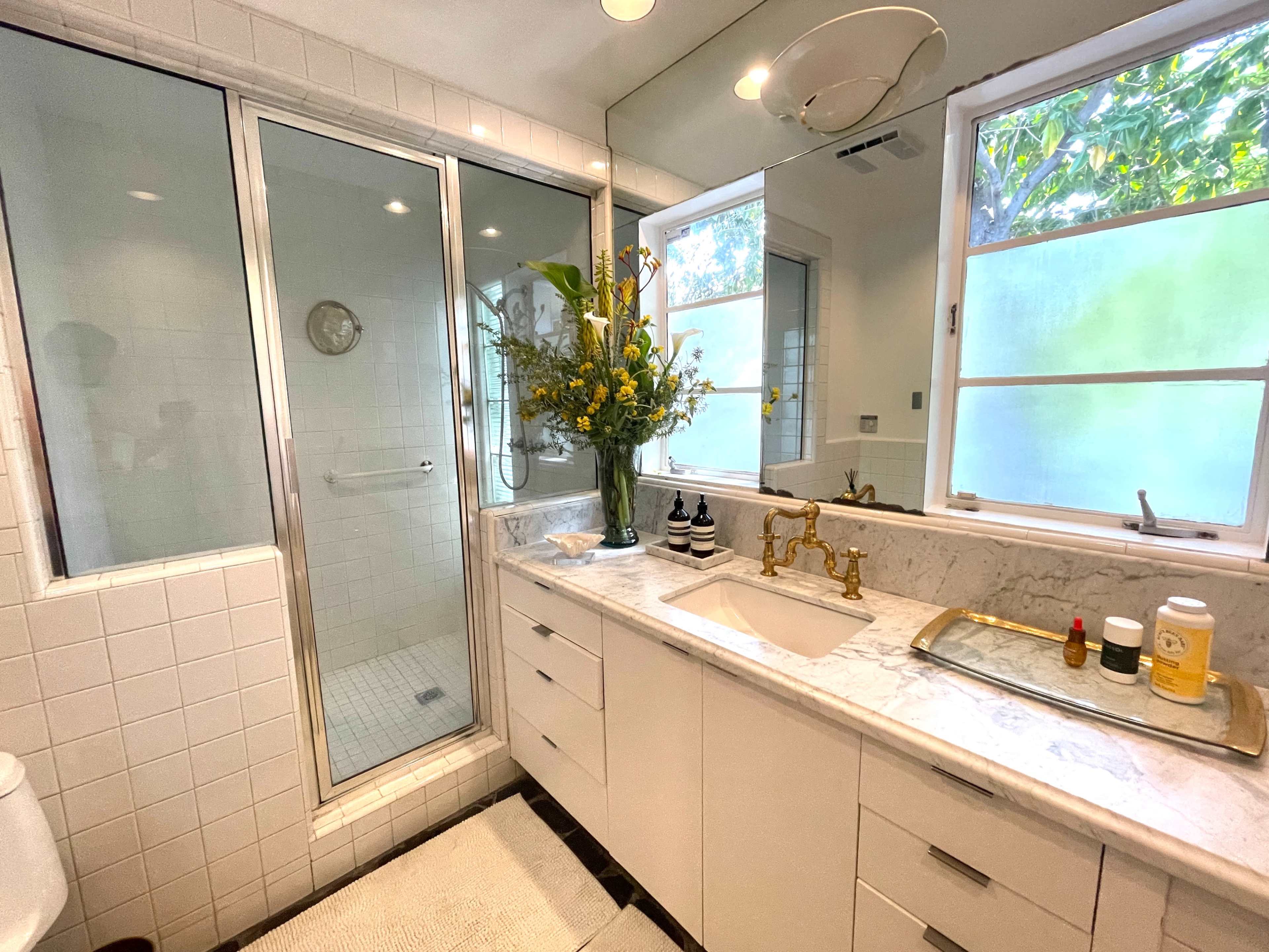 The bathroom features a glass shower enclosure, a marble countertop with a sink, modern fixtures, and large windows that let in natural light.