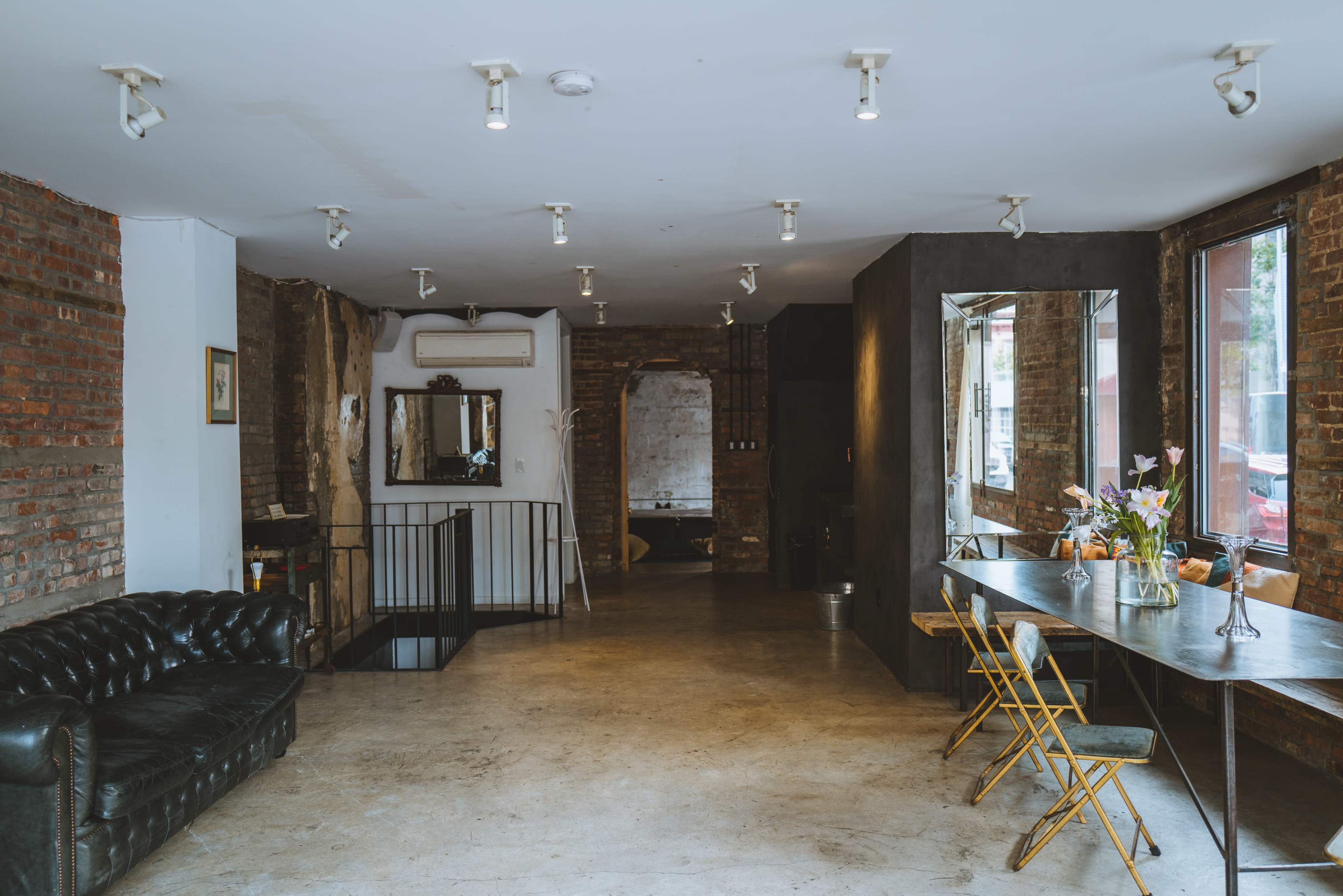 A spacious, minimalist interior with exposed brick walls, a black leather sofa, a metal table with yellow chairs, and large windows.