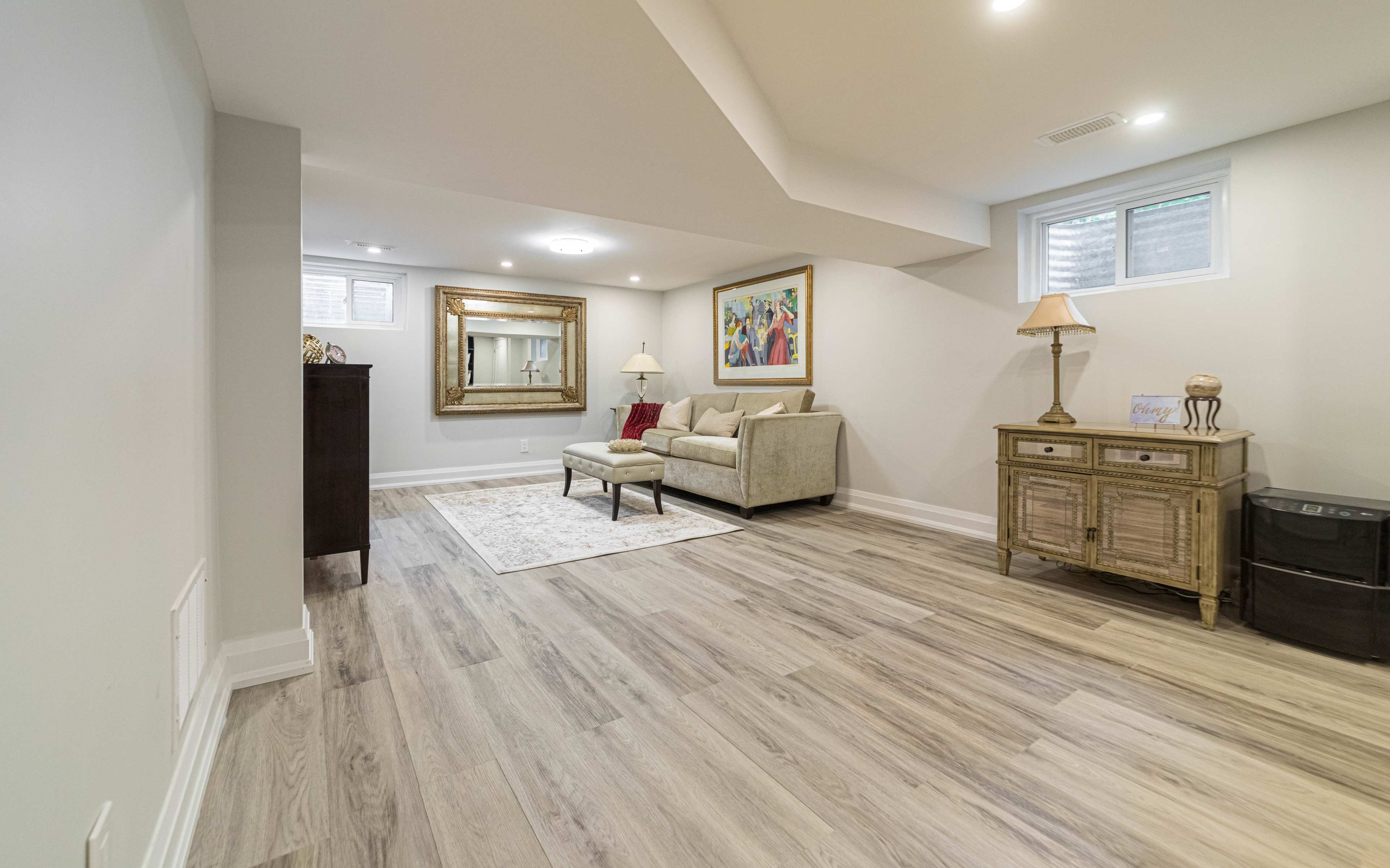 A neatly arranged basement room featuring a light-colored sectional sofa, a decorative mirror, and a wooden side table.