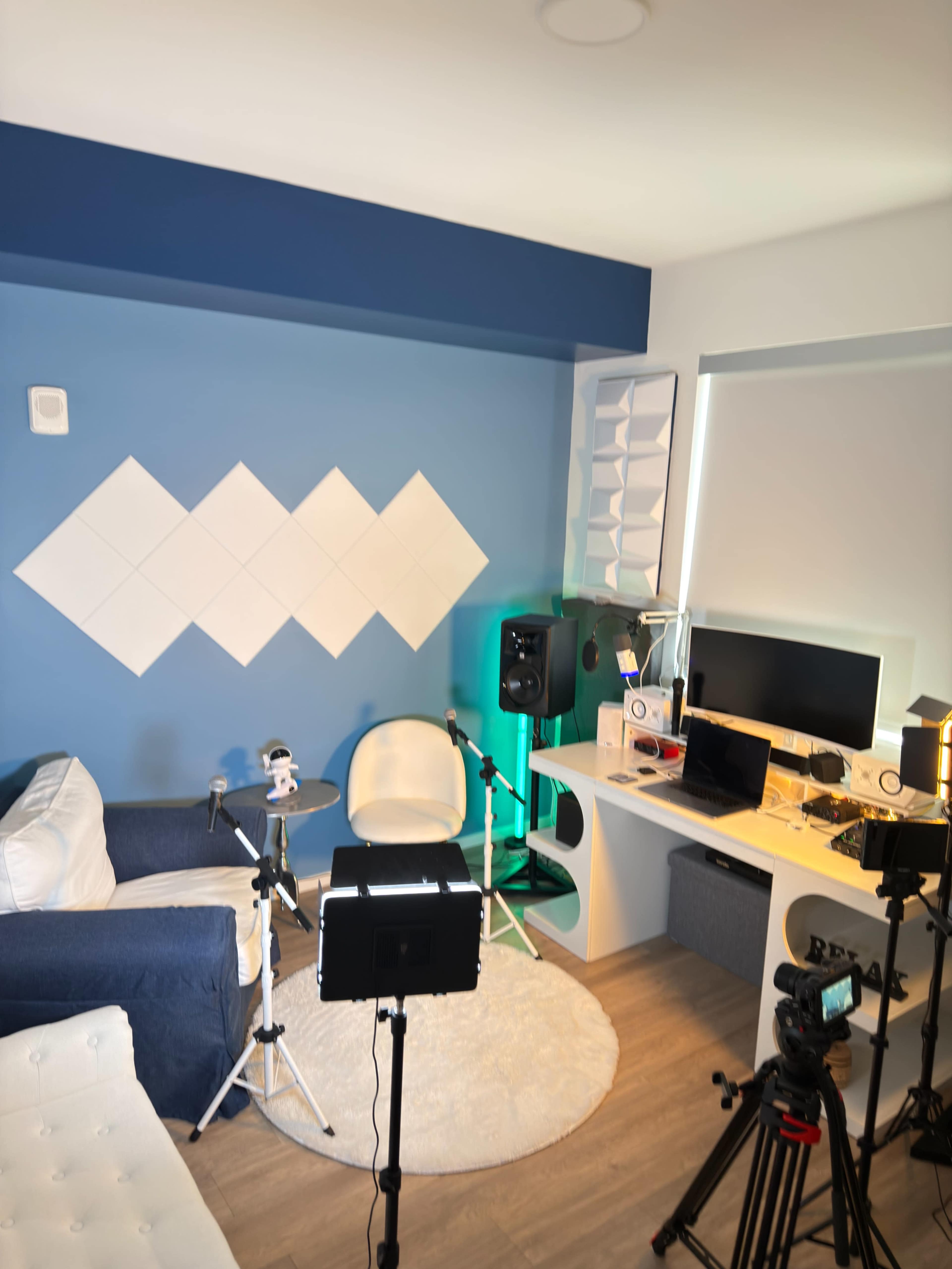 The image shows a bright room with a blue wall featuring decorative diamond shapes, a white couch, a computer setup on a desk, and various equipment for video recording and audio production.