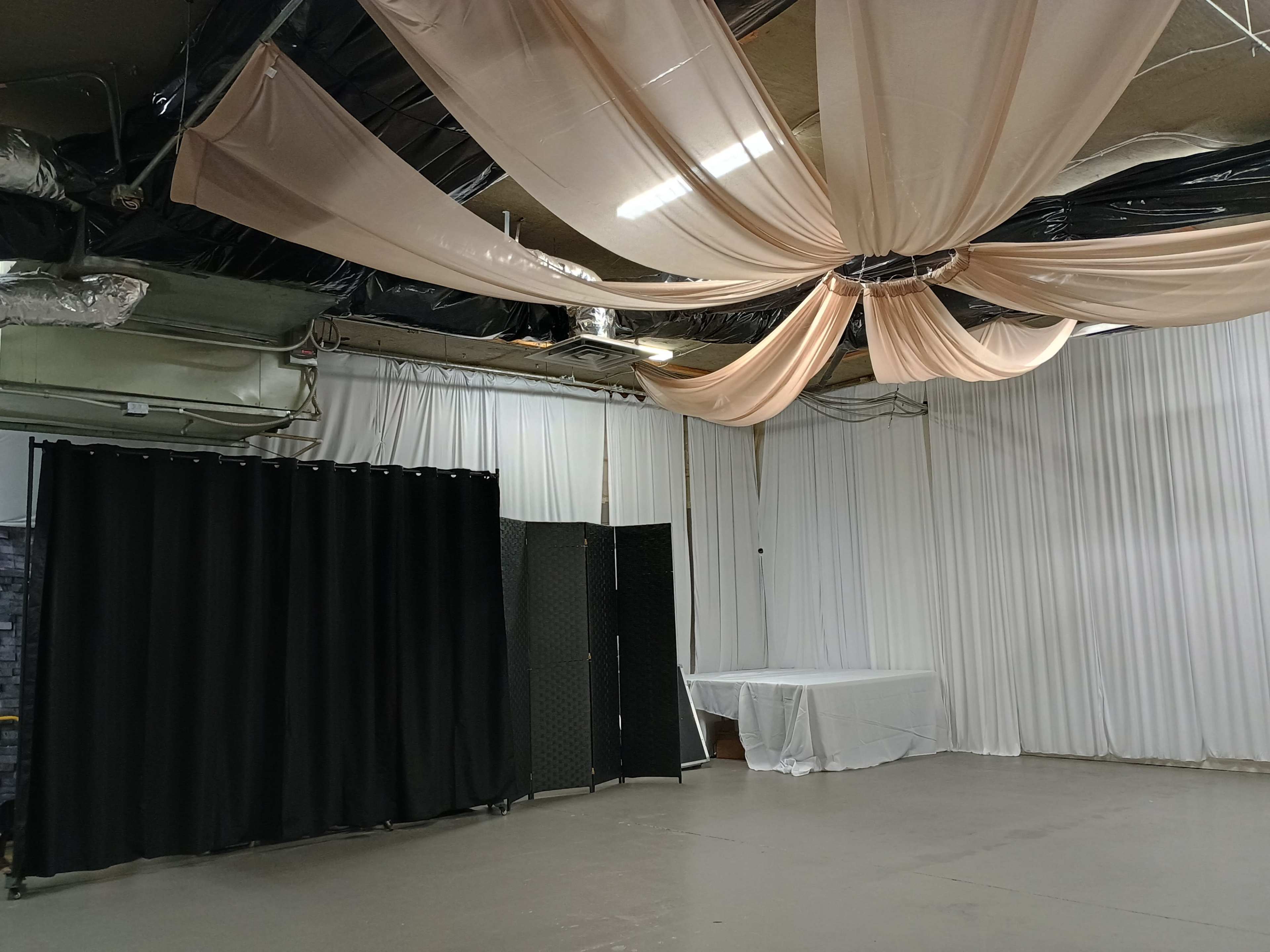 The image shows an indoor space with draped fabric on the ceiling, black curtains on one side, and white drapes covering the walls.