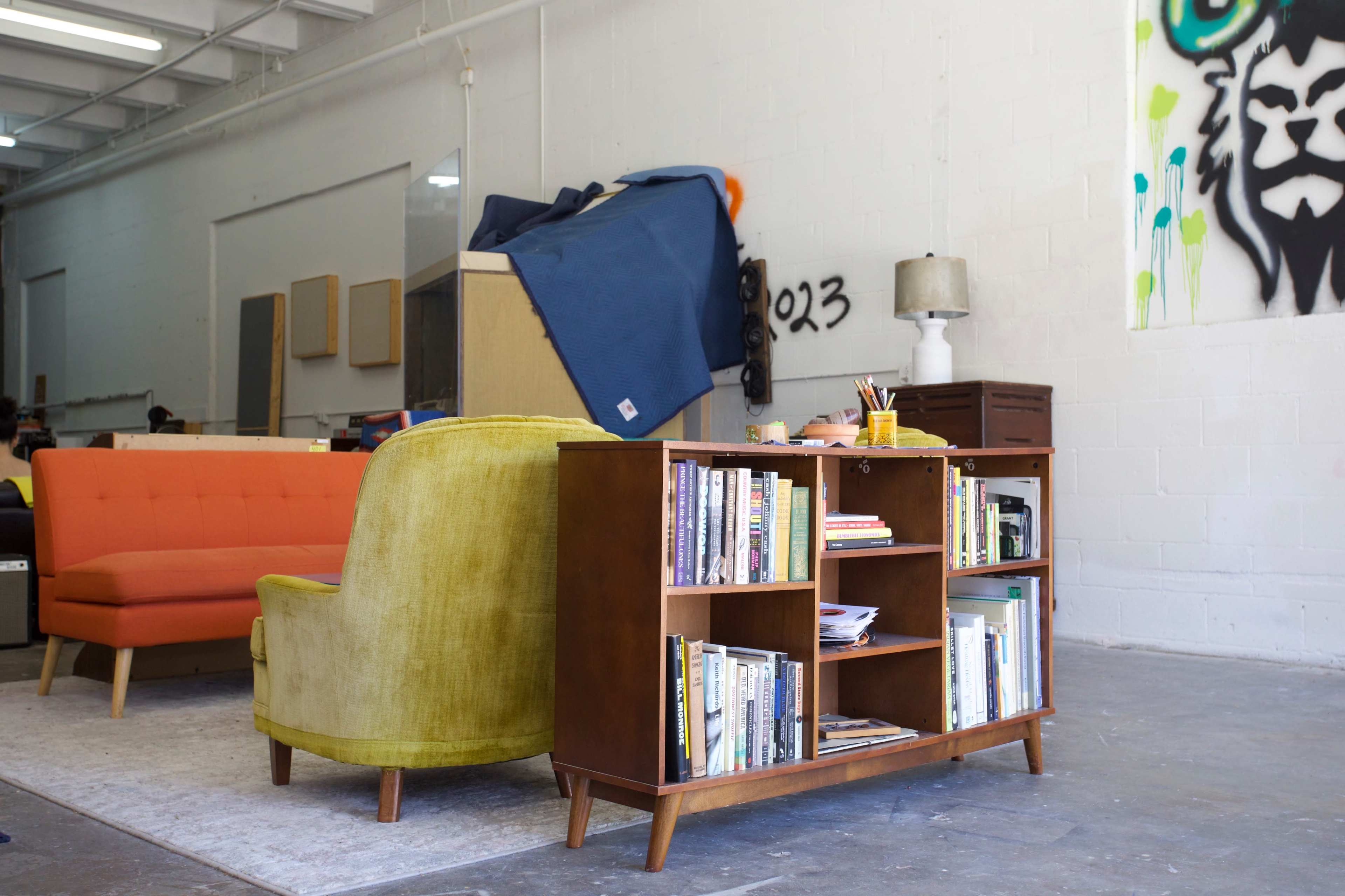 A spacious room with an orange sofa, a green armchair, and a wooden bookshelf filled with books, against a backdrop of white walls and minimal decor.