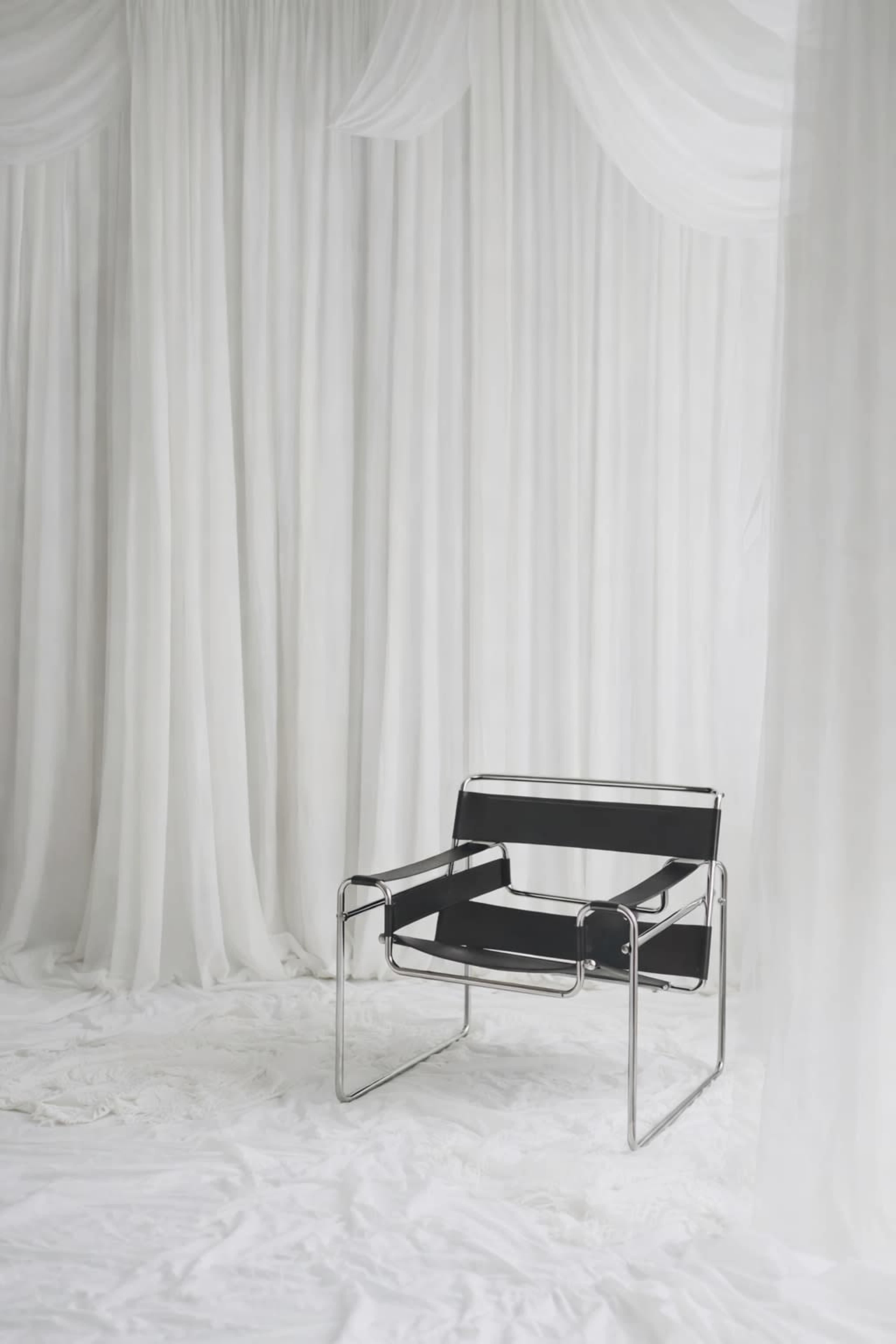 A black metal chair with a minimalist design sits against a backdrop of white curtains and draped fabric.