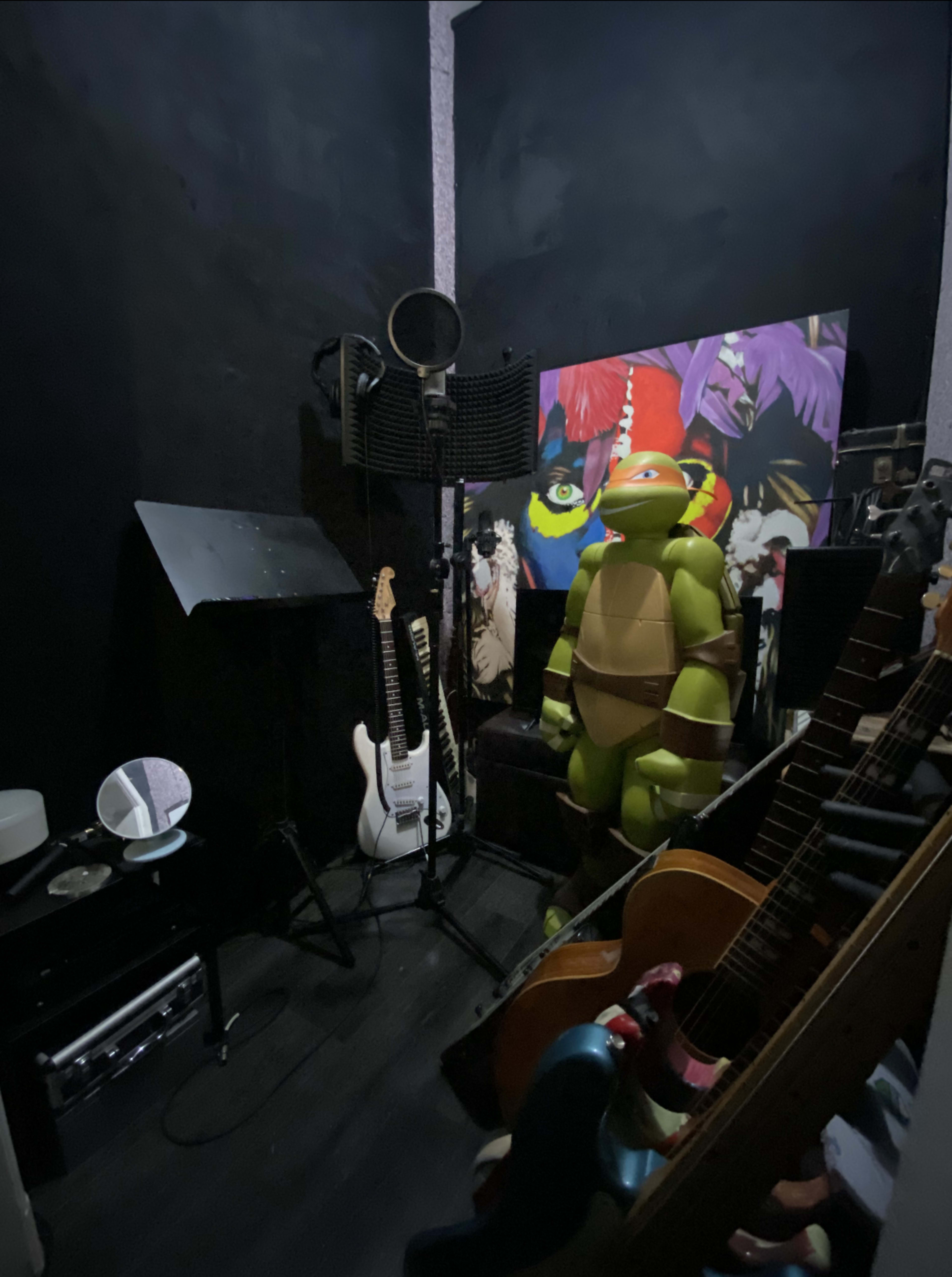 A small music recording room with guitars, a microphone stand, a large poster on the wall, and a life-sized Teenage Mutant Ninja Turtle figure standing among the instruments.