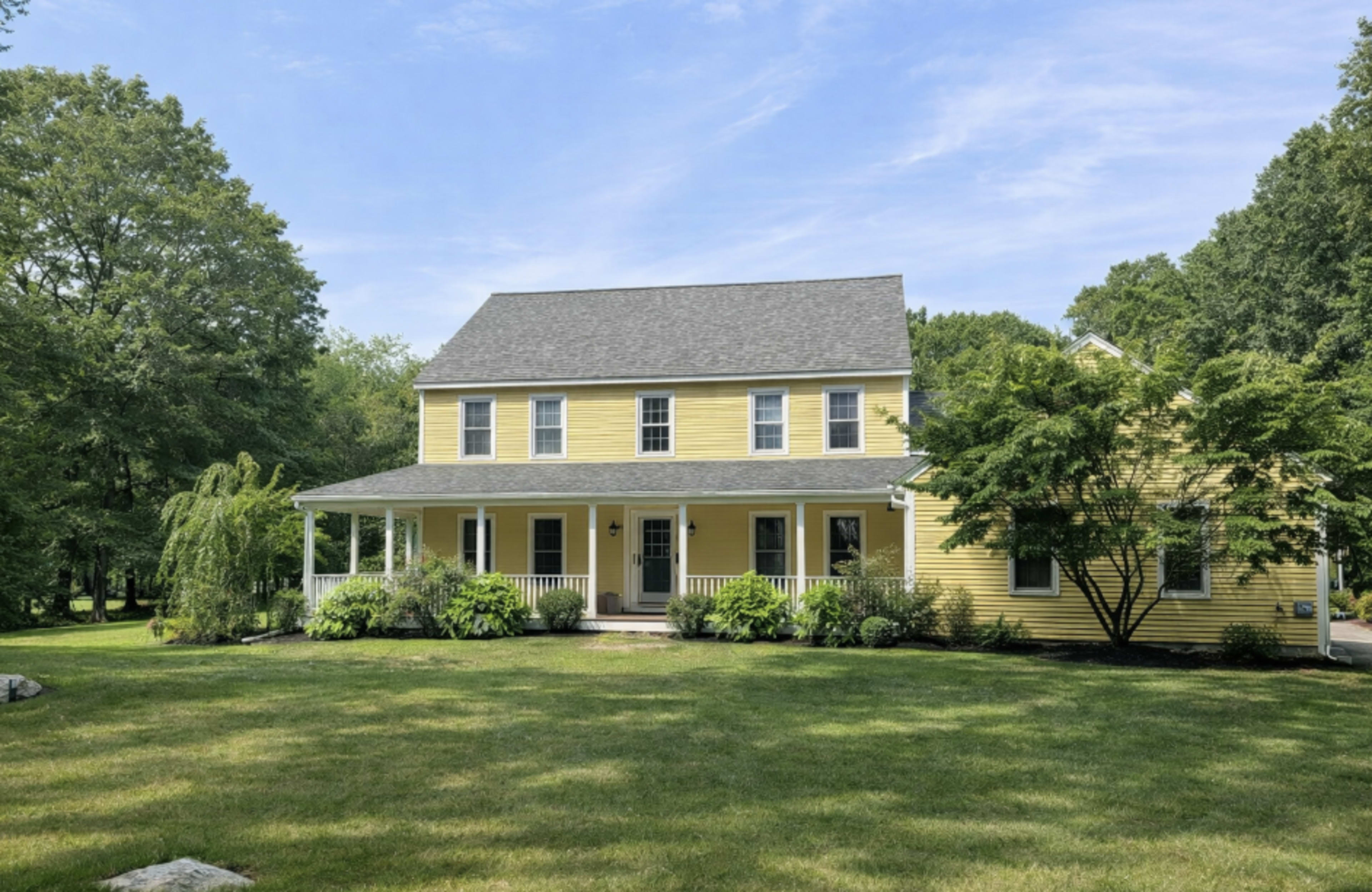 The image shows a two-story yellow house with a front porch, surrounded by greenery and trees.