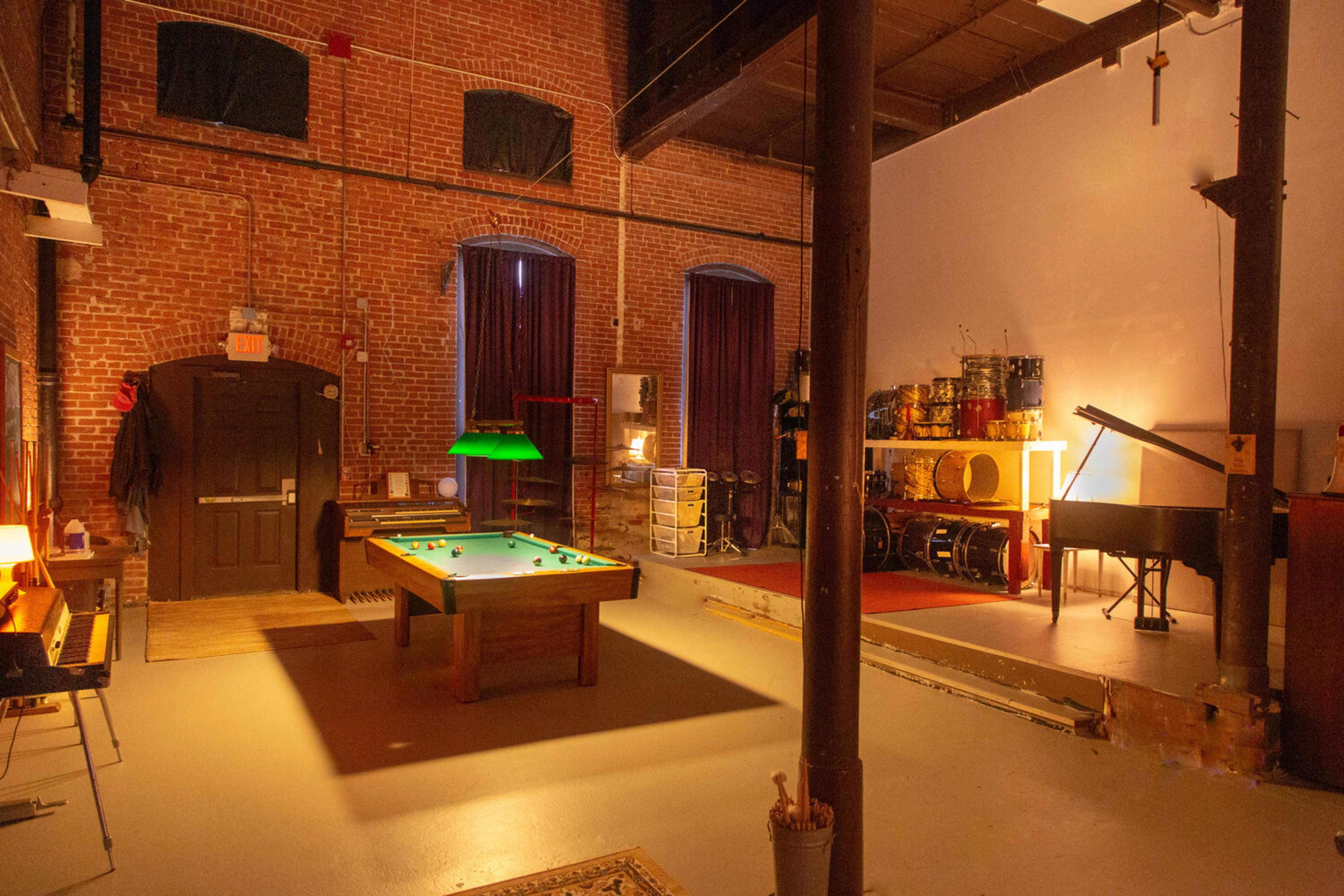 A spacious room with exposed brick walls, featuring a green pool table, a grand piano, and various musical instruments arranged in a corner.