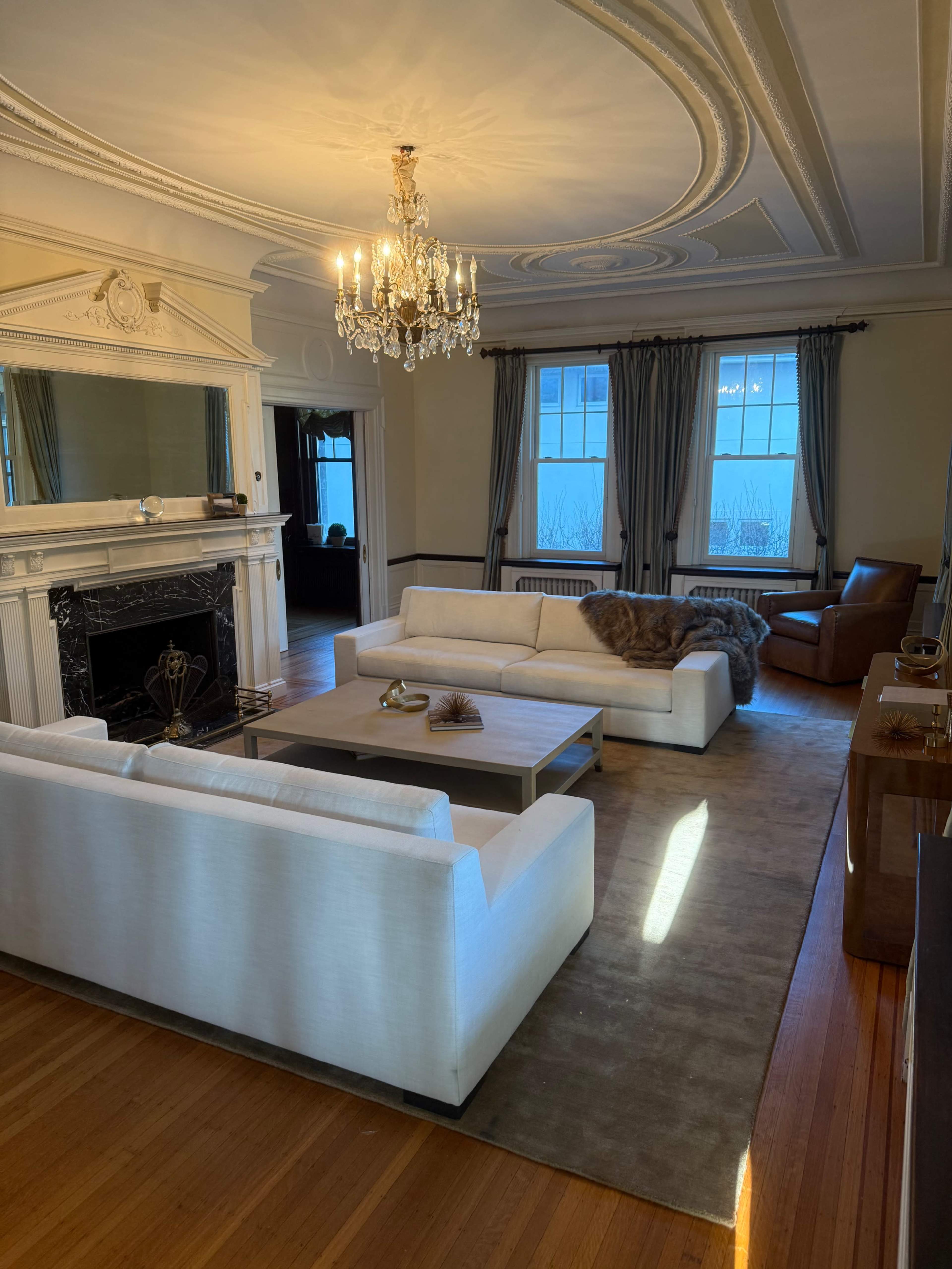 The image shows a living room with a chandelier, a marble fireplace, two white sofas, a brown leather chair, and large windows letting in natural light.
