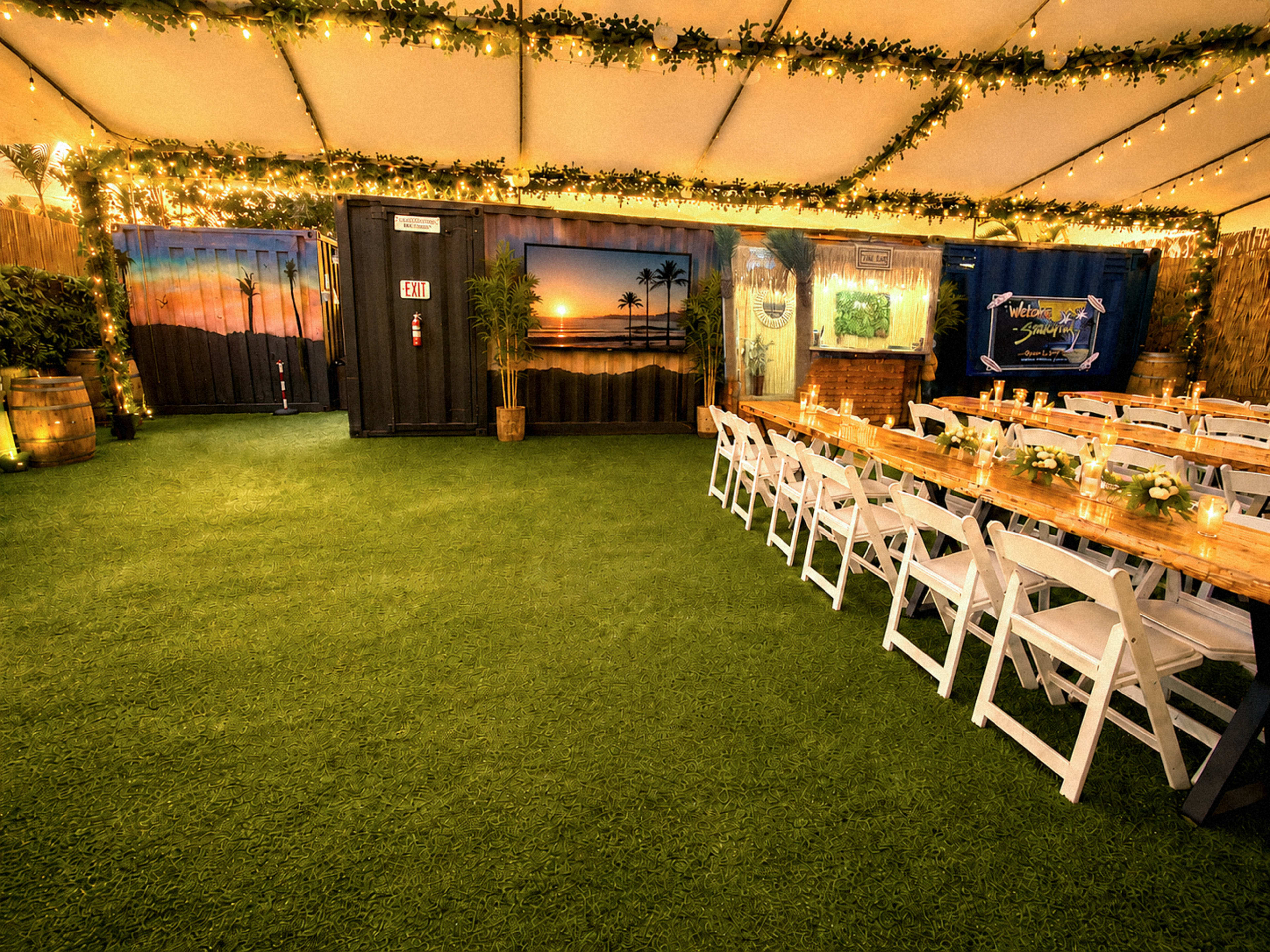 The image shows a decorated indoor event space with tables arranged for dining, artificial grass flooring, and soft lighting from overhead string lights.