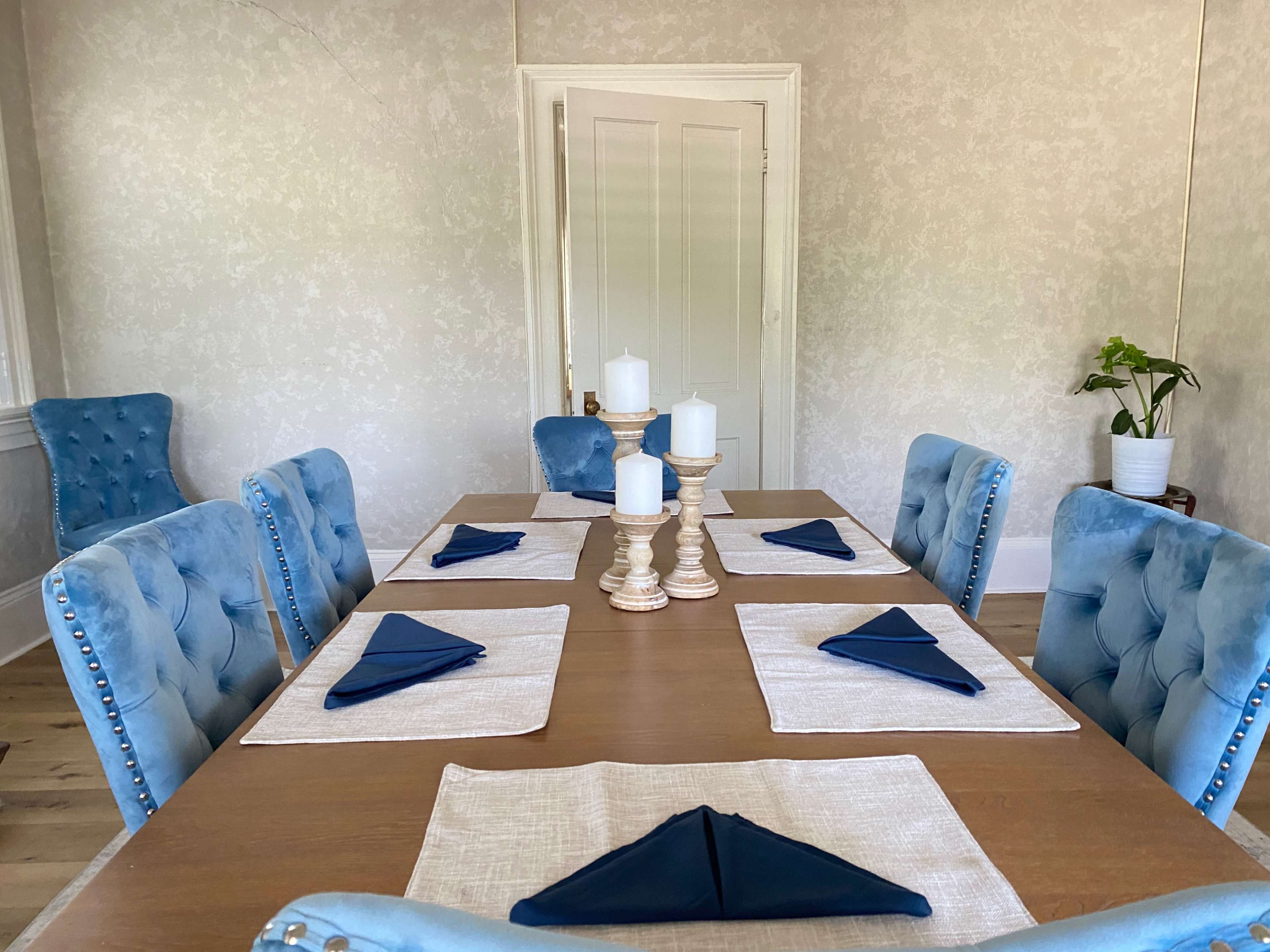 A dining table is set with beige placemats and blue napkins, surrounded by light blue upholstered chairs, with candles at the center and a potted plant in the background.