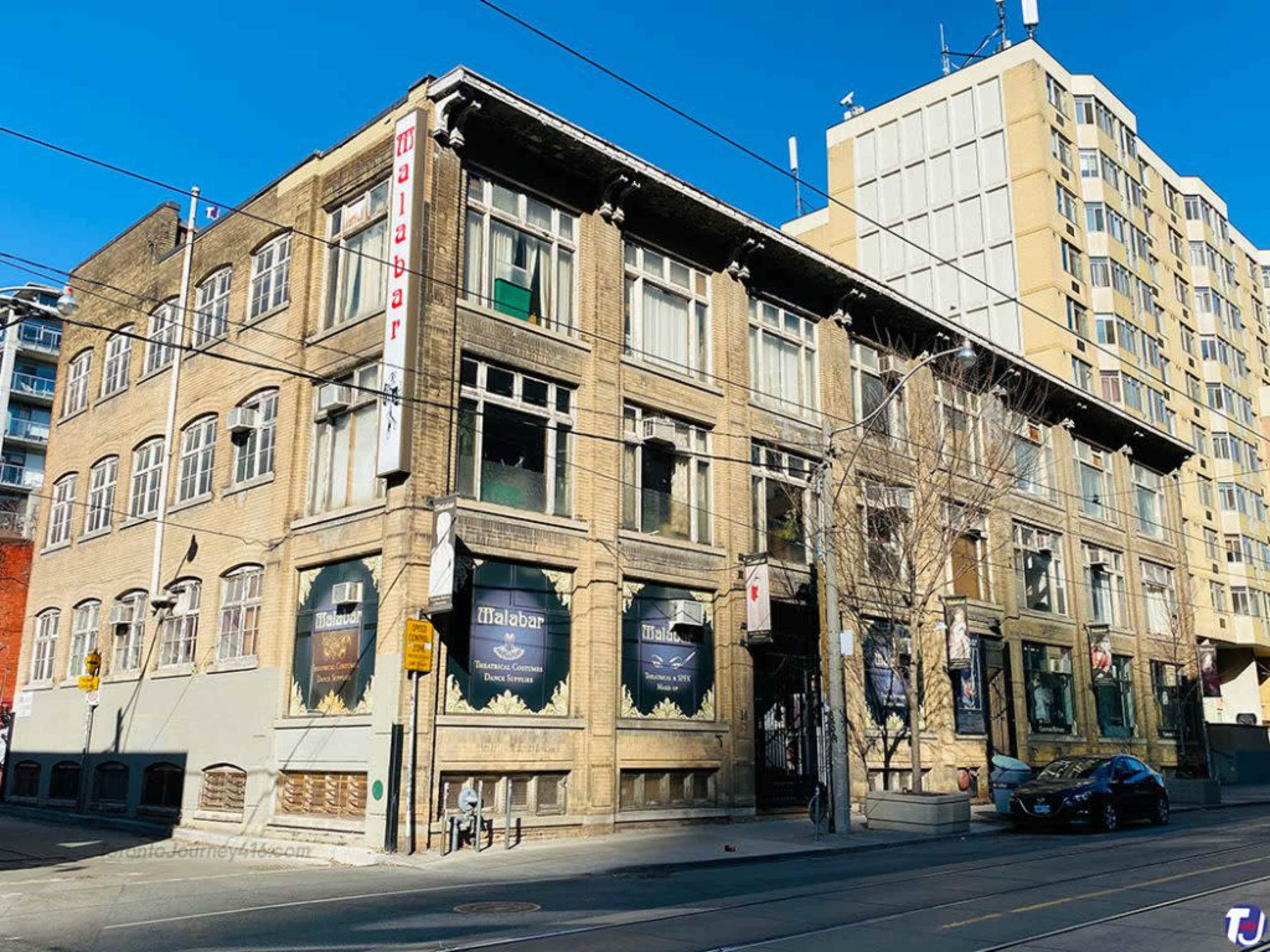 The image shows a historic brick building with large windows, displaying banners and signage, adjacent to a modern high-rise structure.