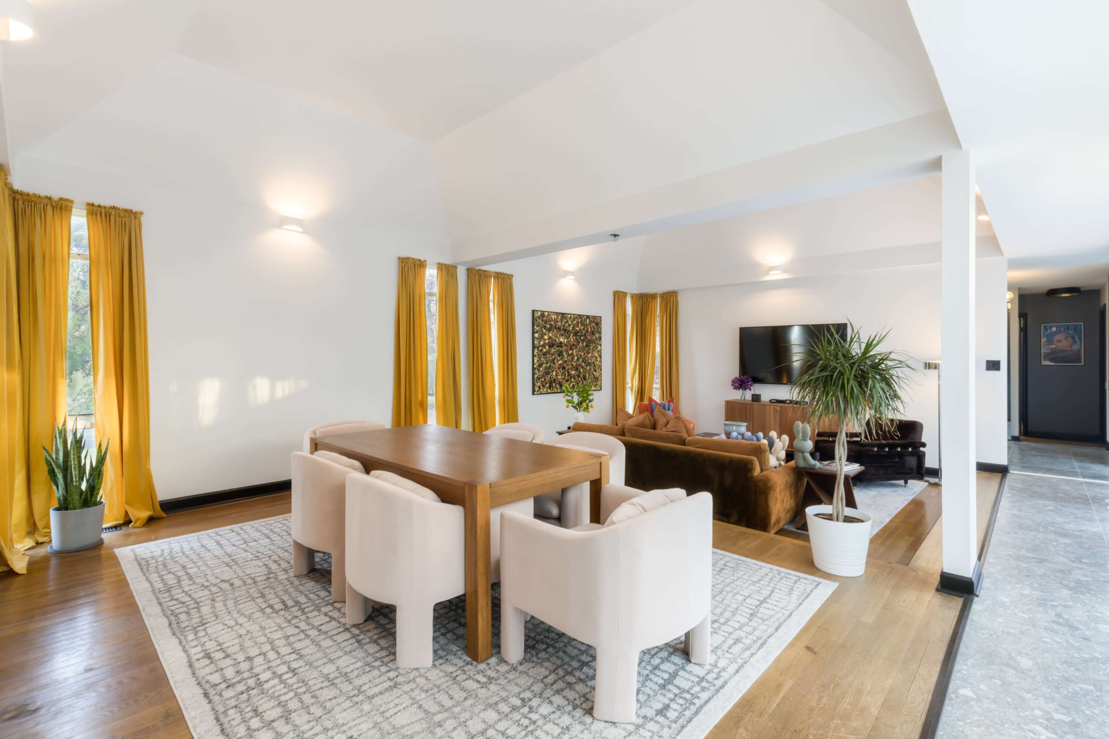 A spacious living and dining area features a wooden table surrounded by upholstered chairs, with large windows dressed in yellow curtains and a television mounted on the wall.