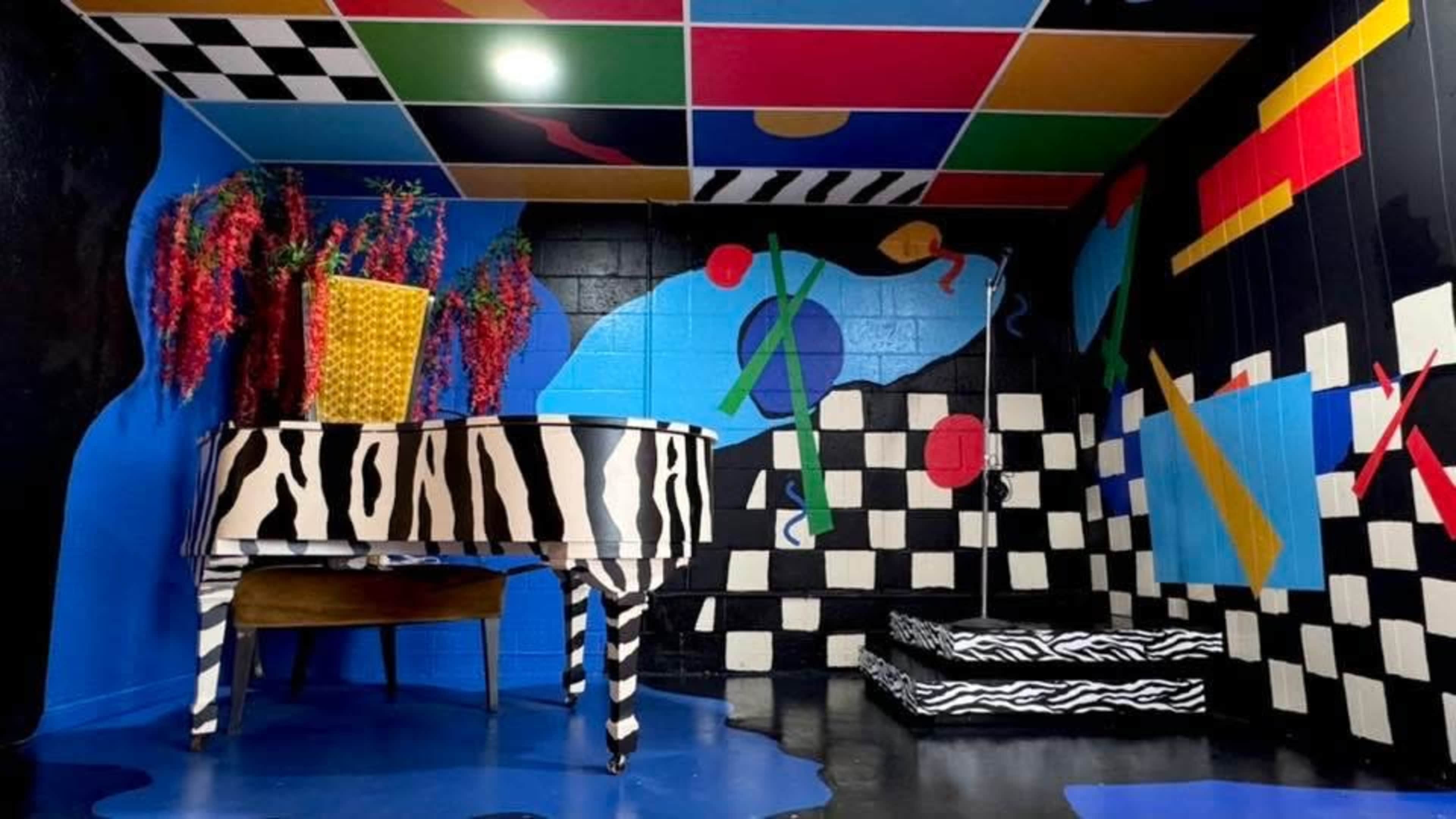 The image shows a colorful room with a zebra-striped piano, geometric wall patterns, and vibrant decor, including a large potted plant.