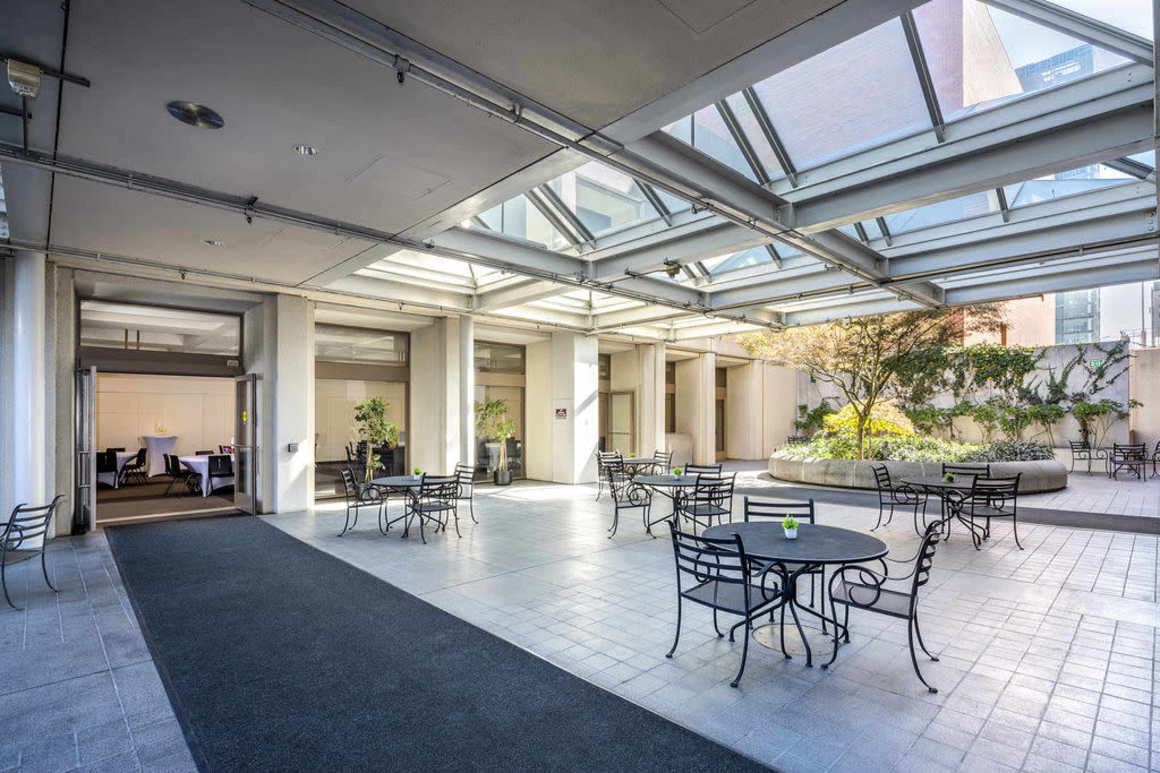 The image shows a modern, open-air courtyard with multiple metal tables and chairs arranged around a central planter filled with greenery.