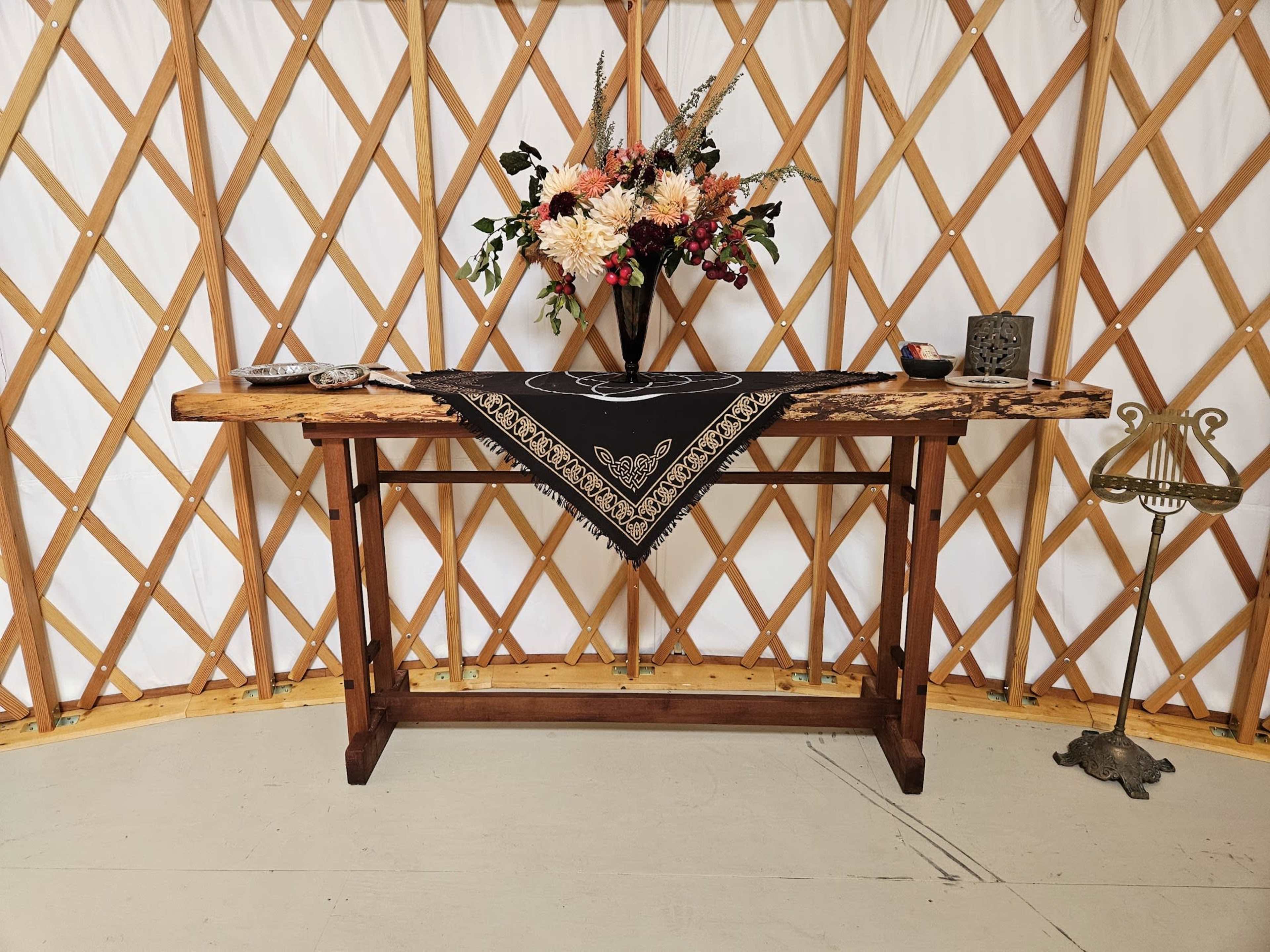 A wooden table with a black cloth and decorative items is positioned inside a circular yurt with a floral arrangement on top.