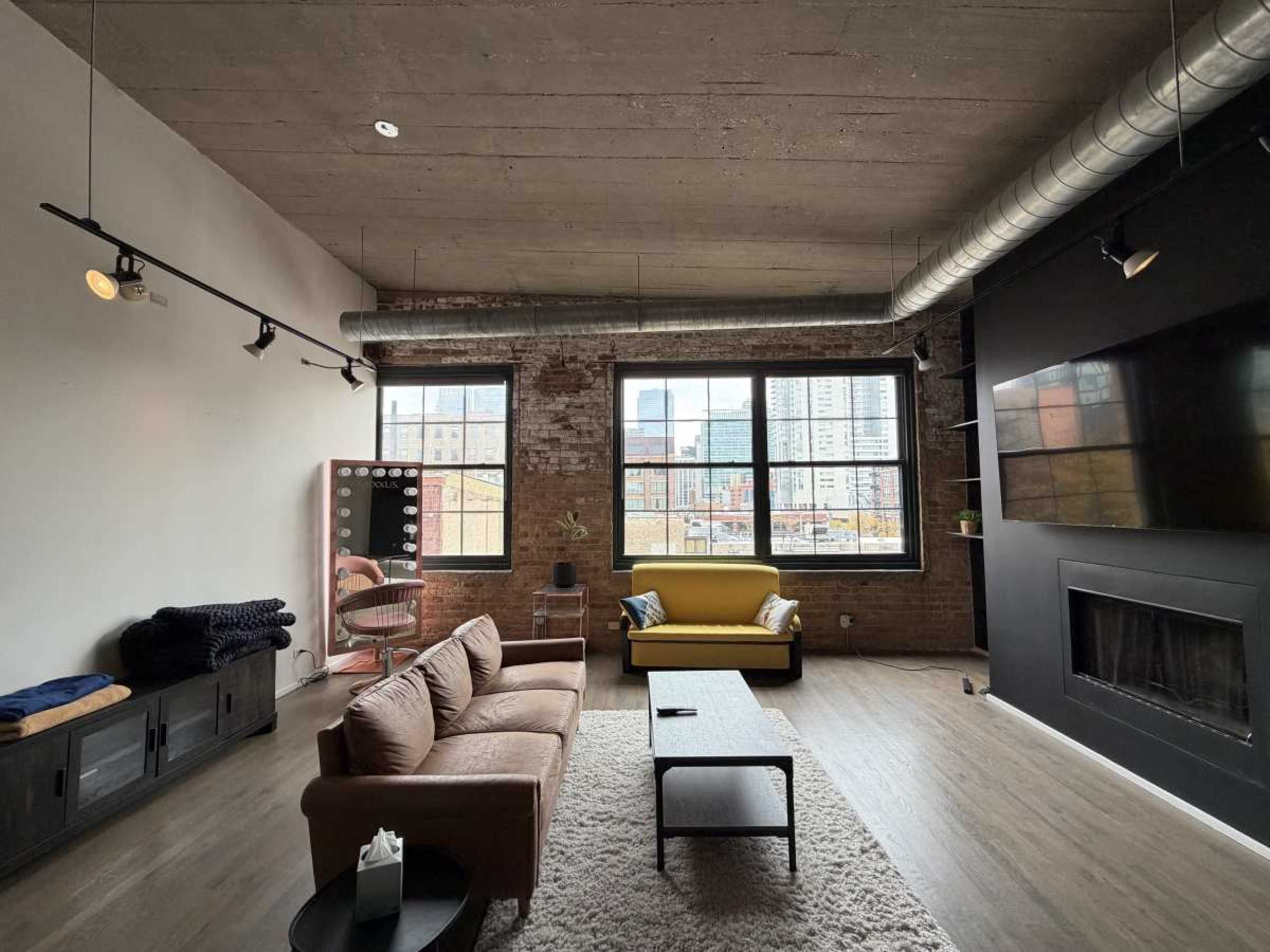 A modern living room features a brown sofa, a yellow armchair, and large windows that showcase a city view, with exposed brick walls and concrete ceiling.