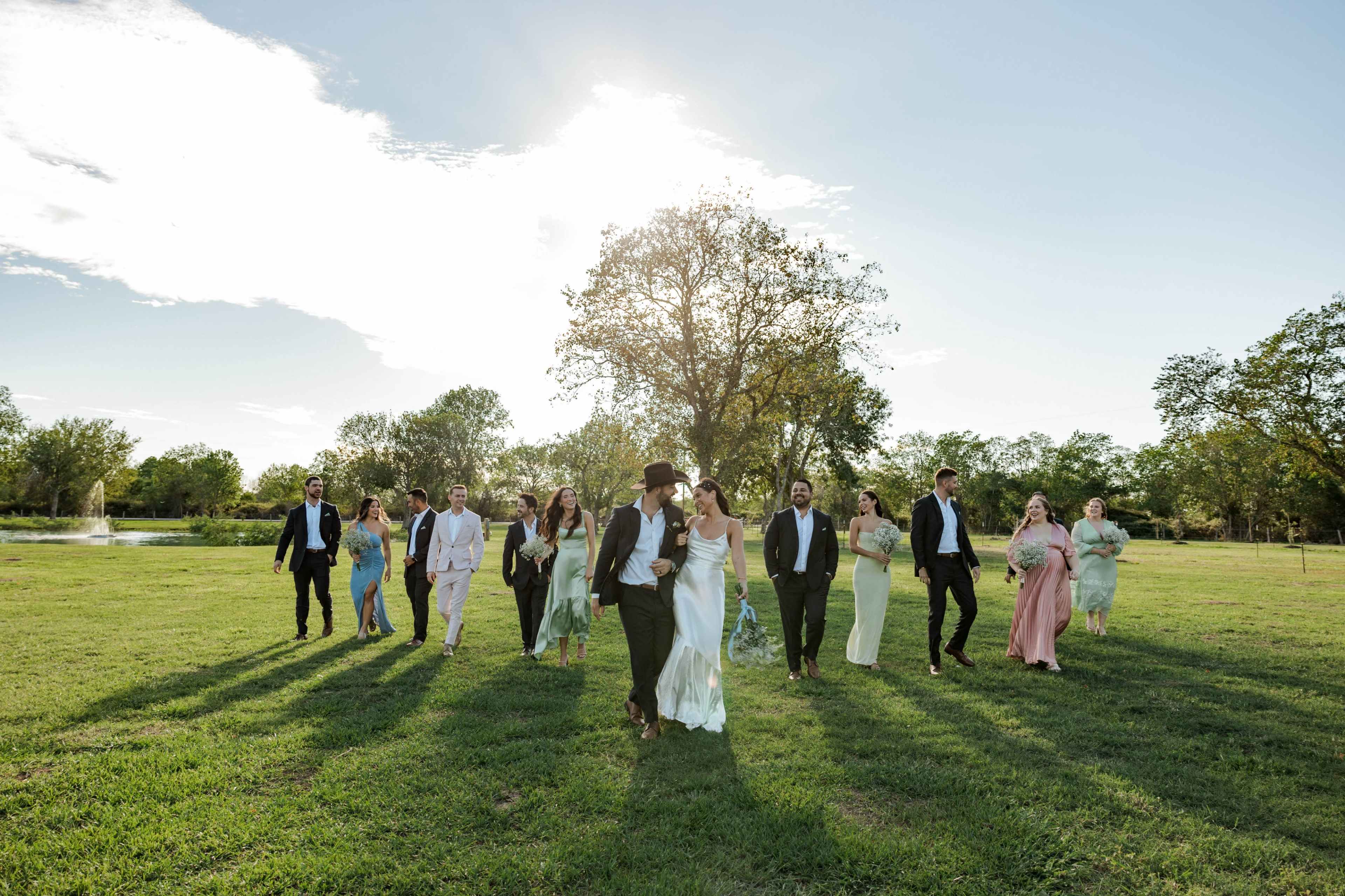 A group of people dressed in formal attire walks across a grassy field with a pond visible in the background.