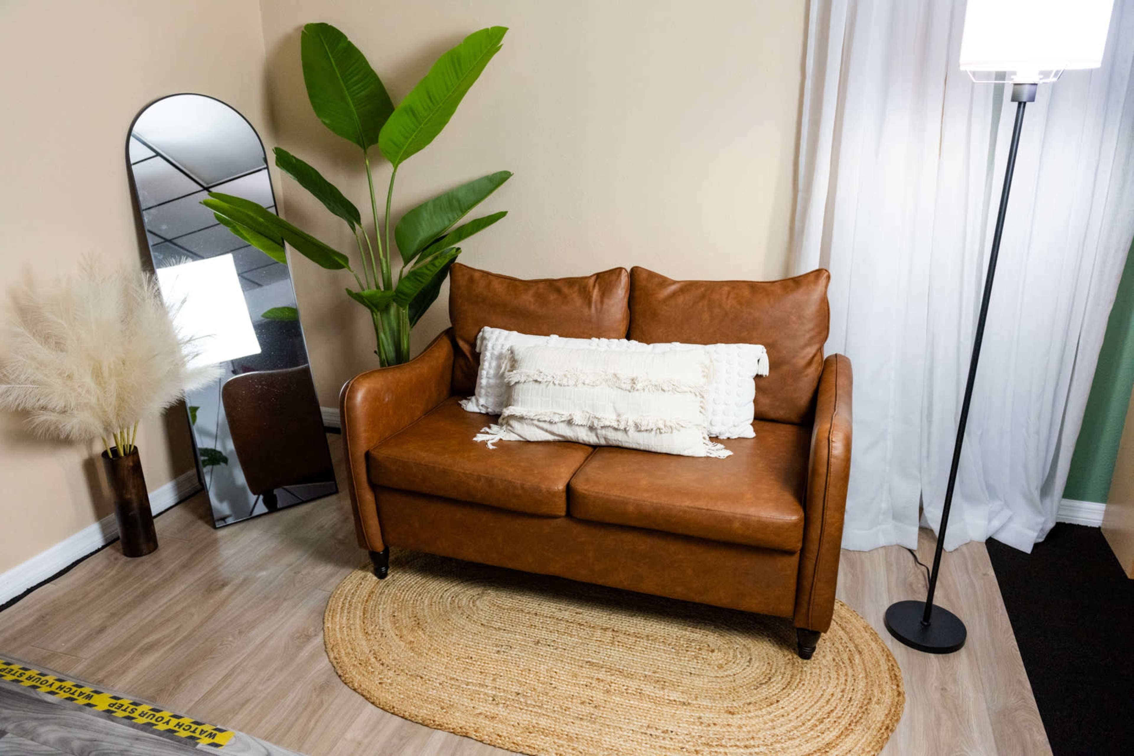 A brown leather sofa sits on a round rug in a well-lit room adorned with a tall plant, a mirror, and a floor lamp.