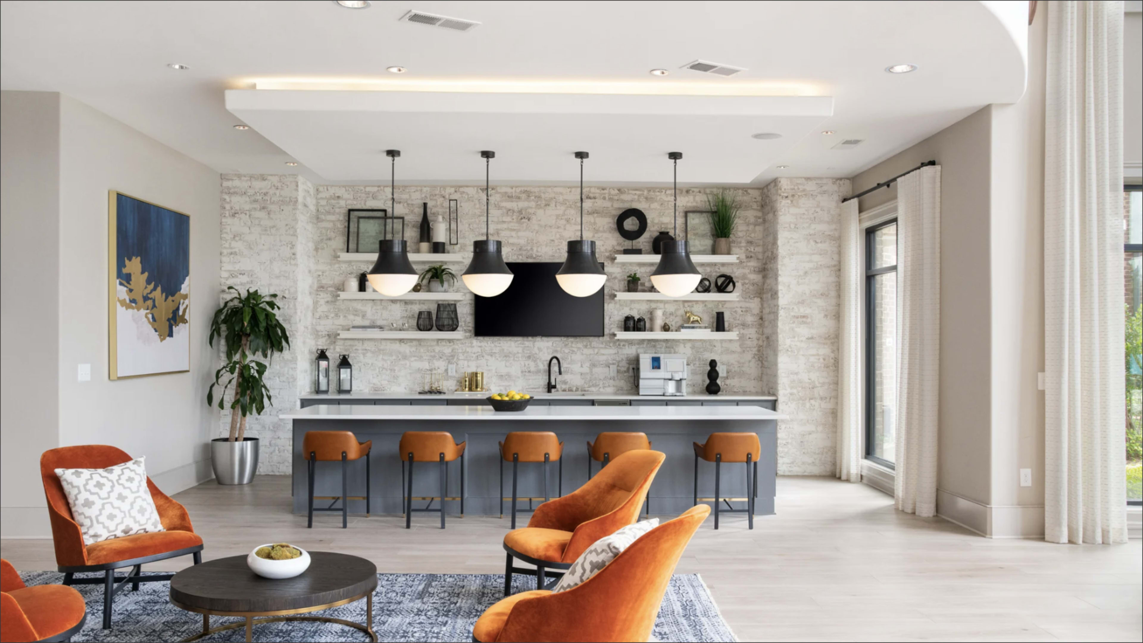 A modern living area features a kitchen bar with gray cabinetry and orange chairs, pendant lights hanging above, and a decorative wall with plants and artwork.
