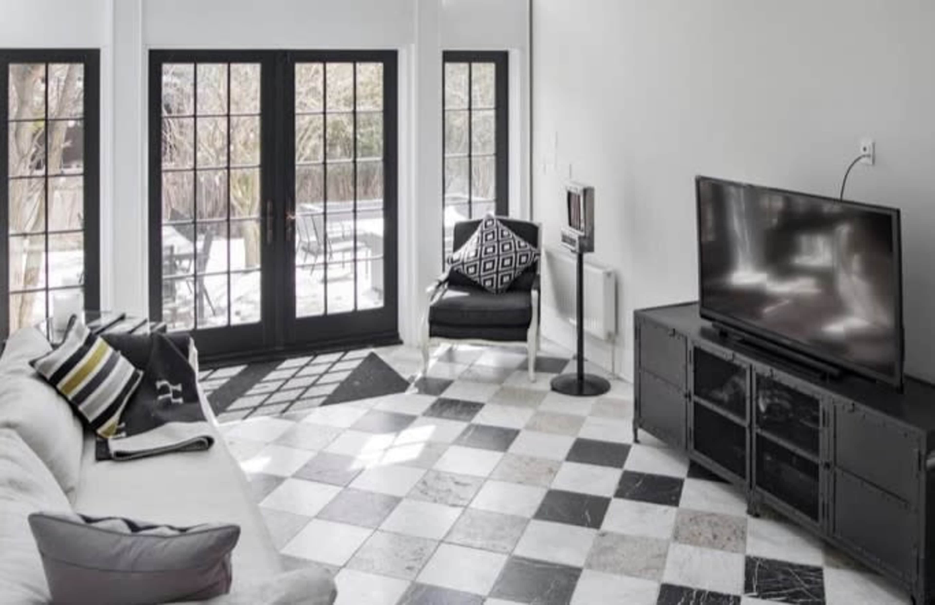 A modern living room with a black-and-white checkered floor, a gray sofa, and a television facing a set of glass doors leading to an outdoor space.