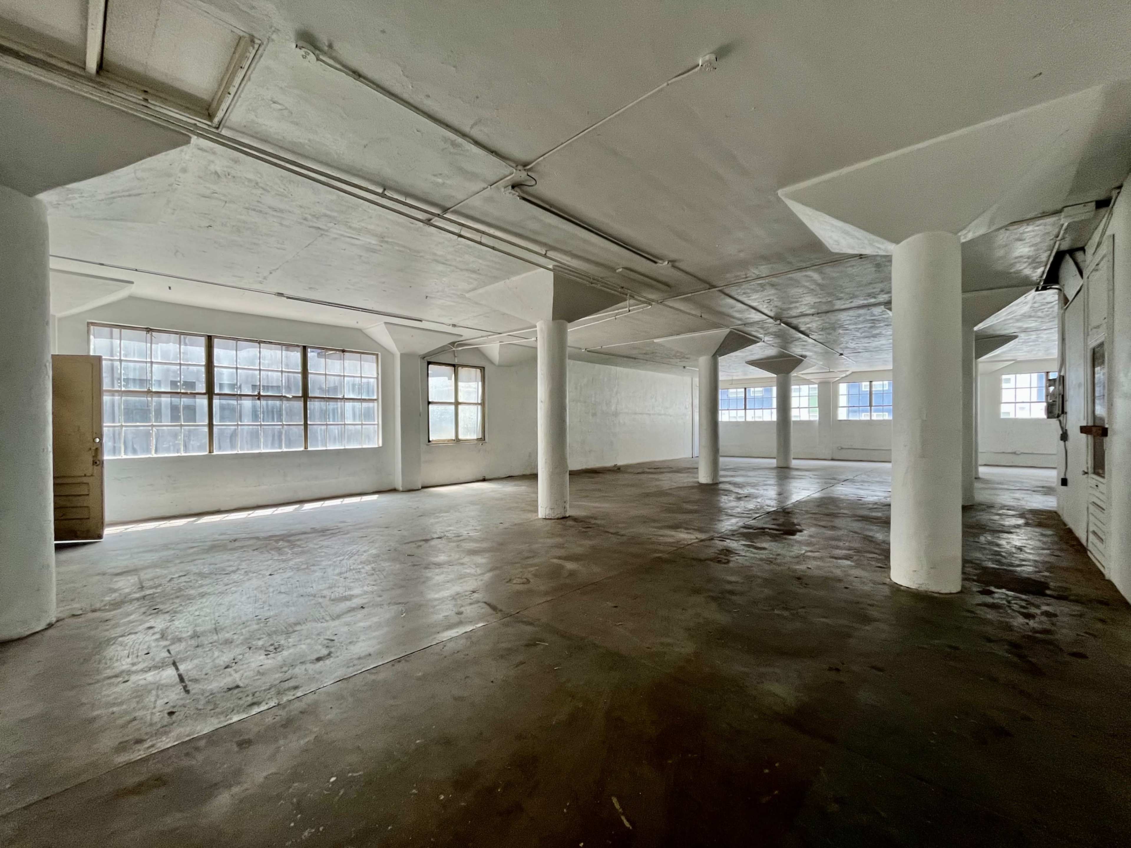 The image shows a spacious, empty interior of a building with concrete pillars and large windows letting in natural light.