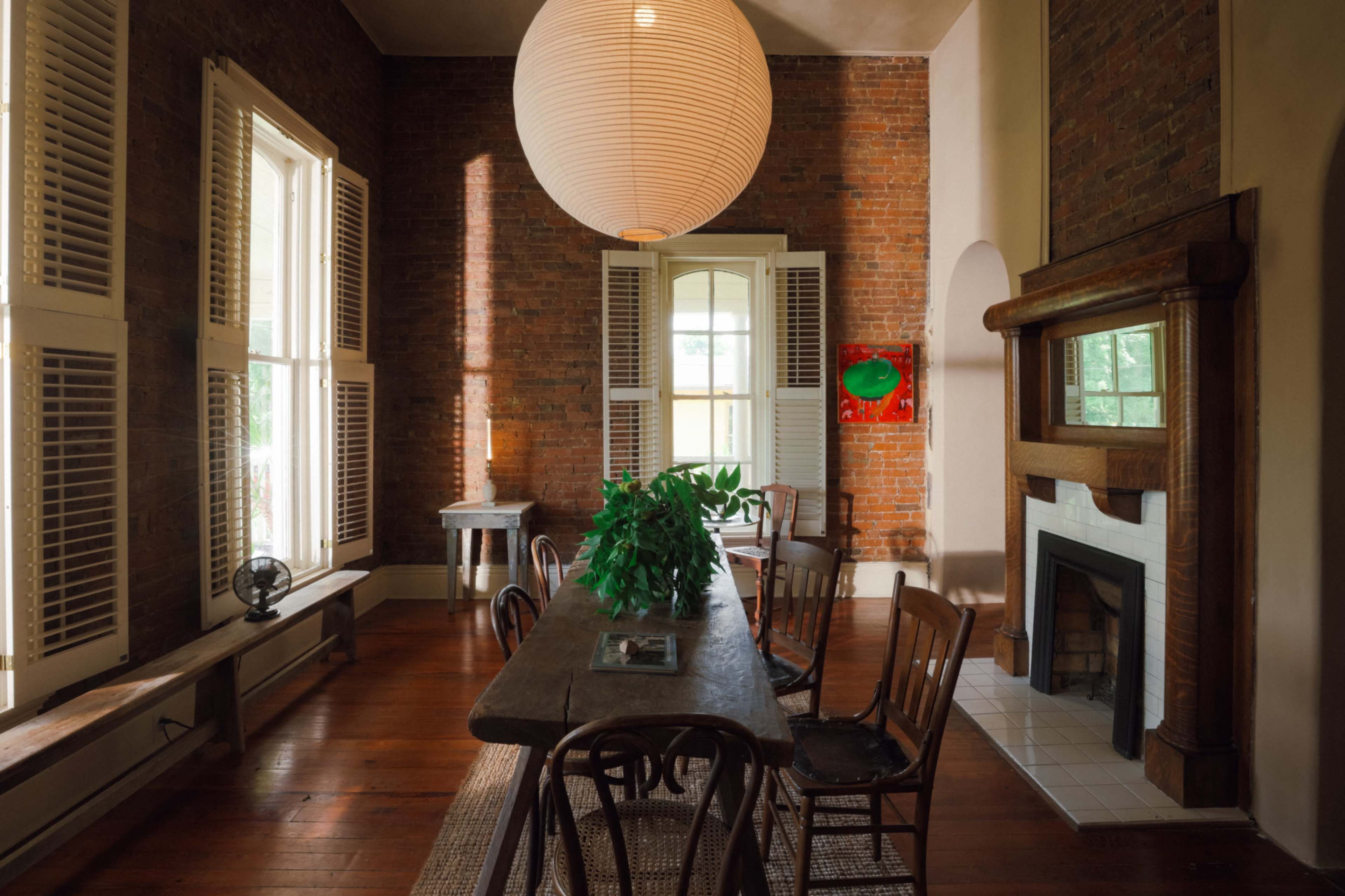 A dining area features a wooden table surrounded by chairs, illuminated by natural light through large windows and a hanging pendant lamp, with exposed brick walls and a fireplace.