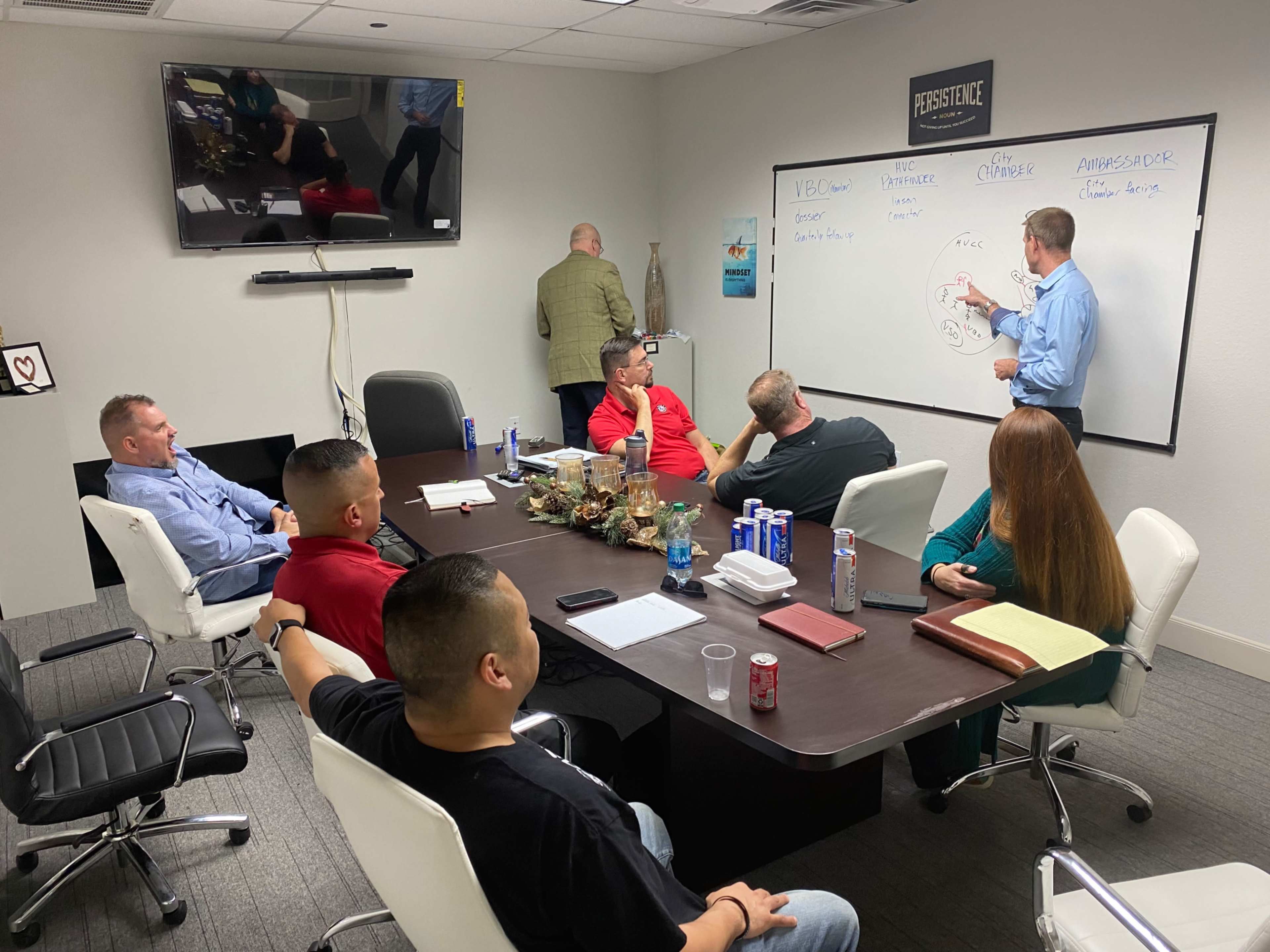 A group of seven people is seated around a conference table, while one person stands and writes on a whiteboard in a meeting room.