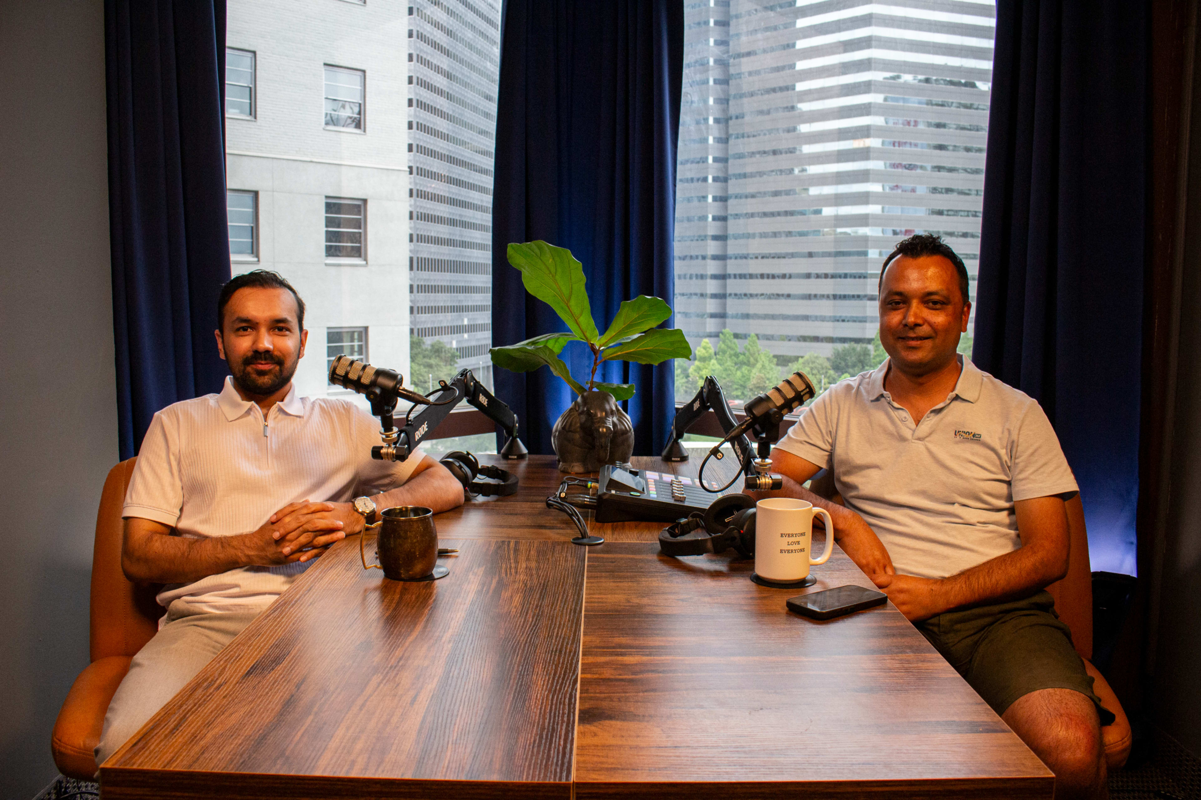 Two men sit at a wooden table in a studio with microphones, a plant centerpiece, and large windows showing a cityscape in the background.