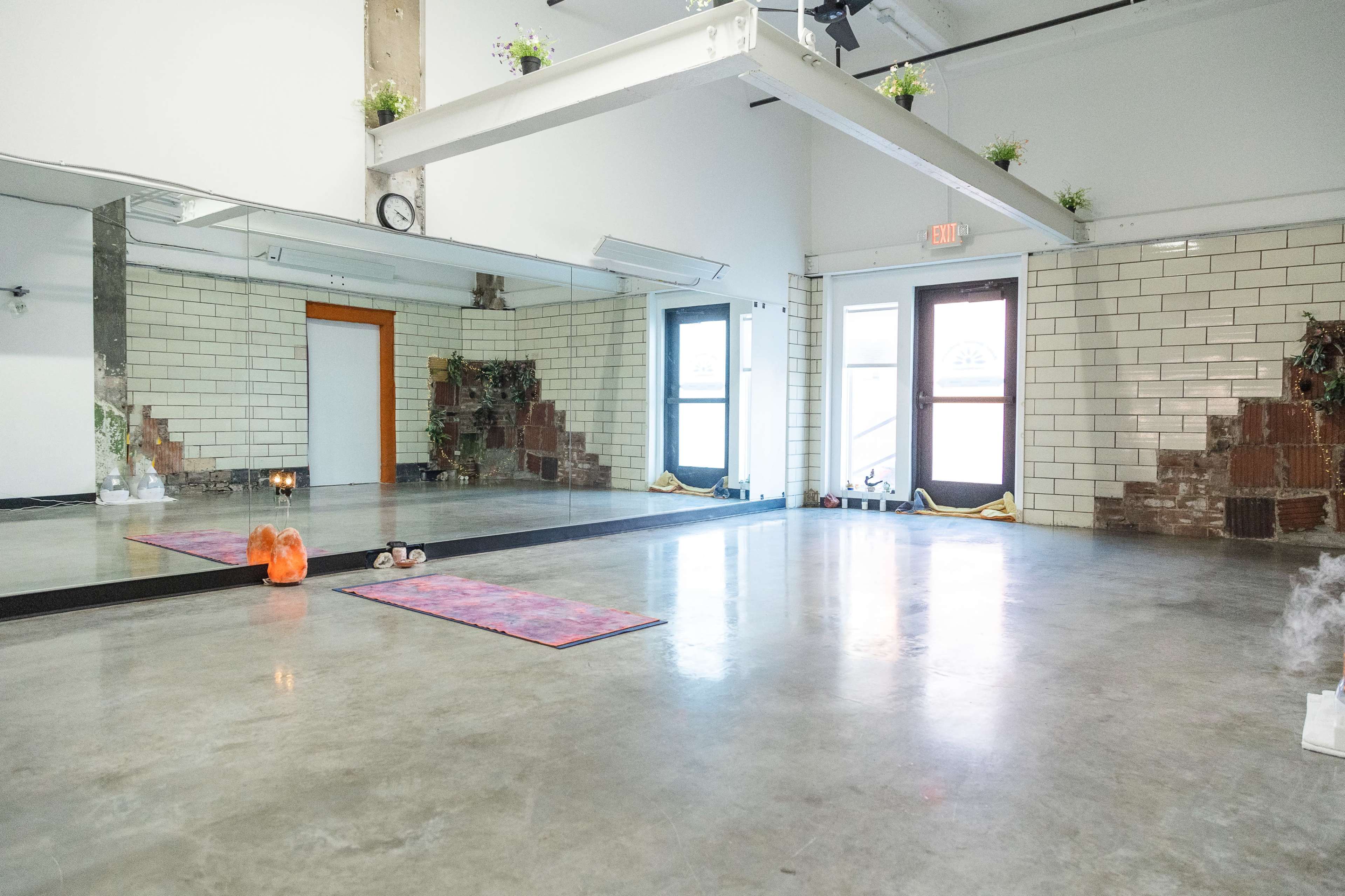 A spacious, empty yoga studio with a mirror and yoga mat on the floor, featuring brick walls and large windows.