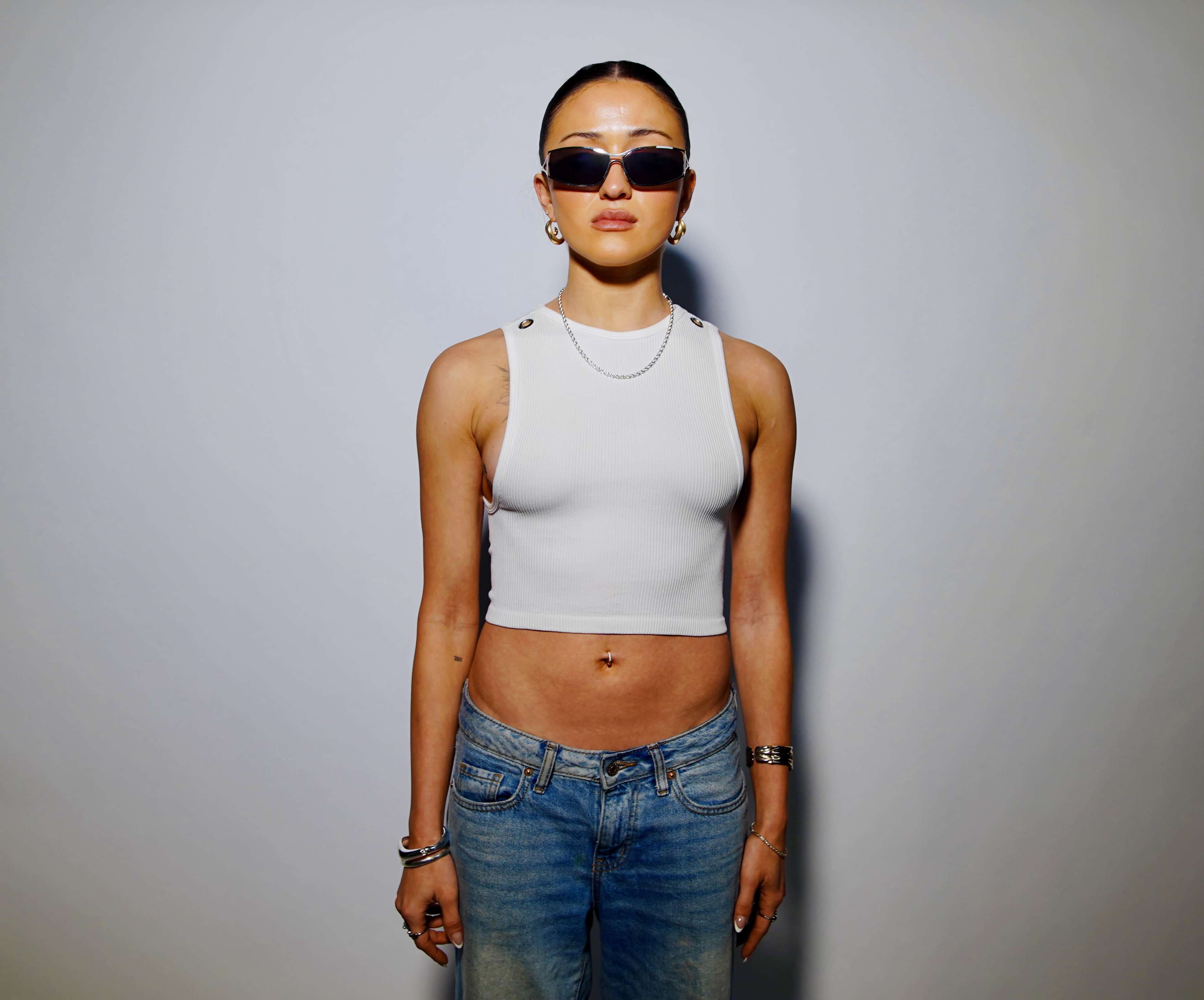 A person stands against a plain background, wearing a sleeveless white top and blue jeans, with sunglasses and visible jewelry.