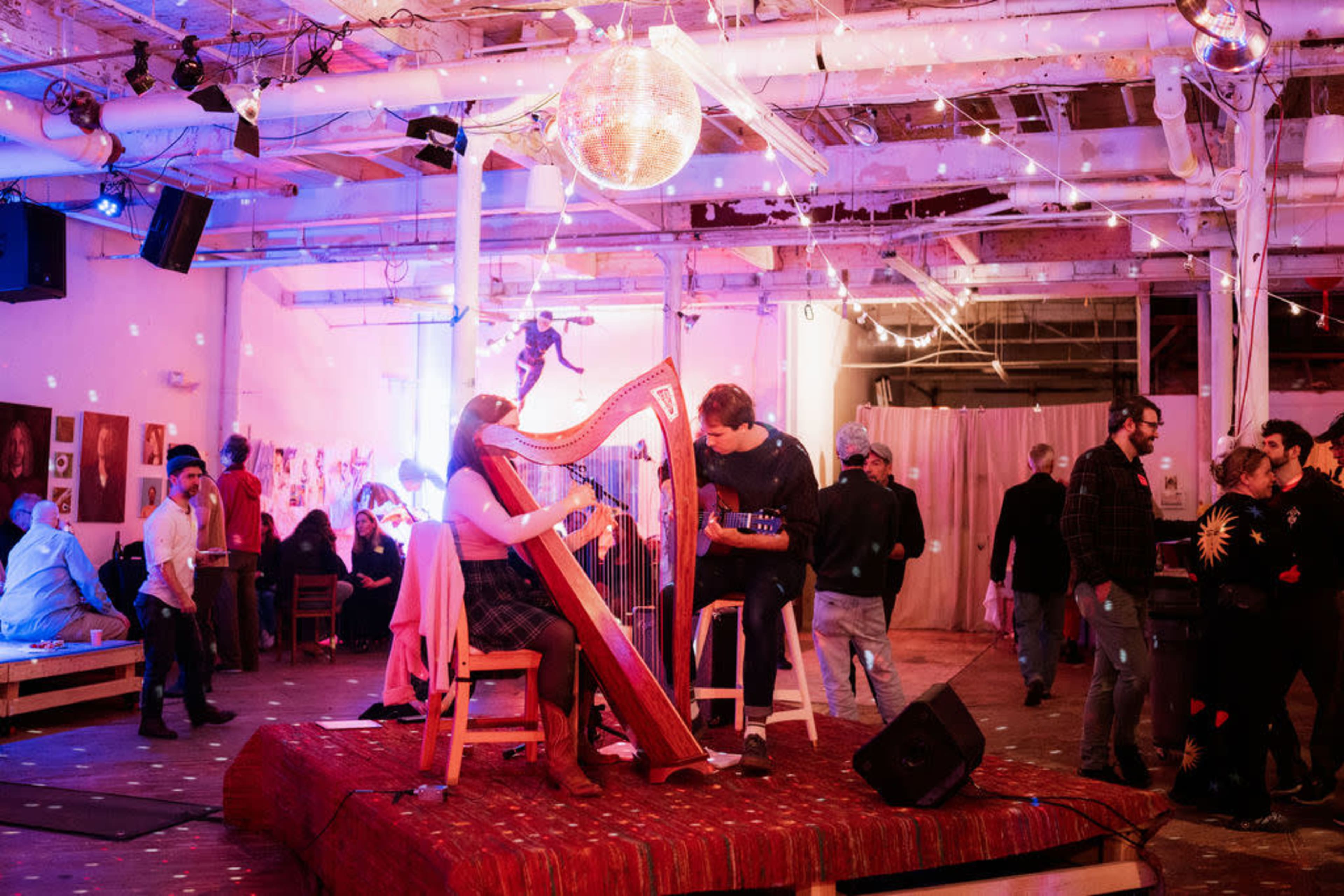 A musician plays a harp while another strums a guitar on a stage in a lively art space, with people mingling and colorful lights reflecting off a disco ball.