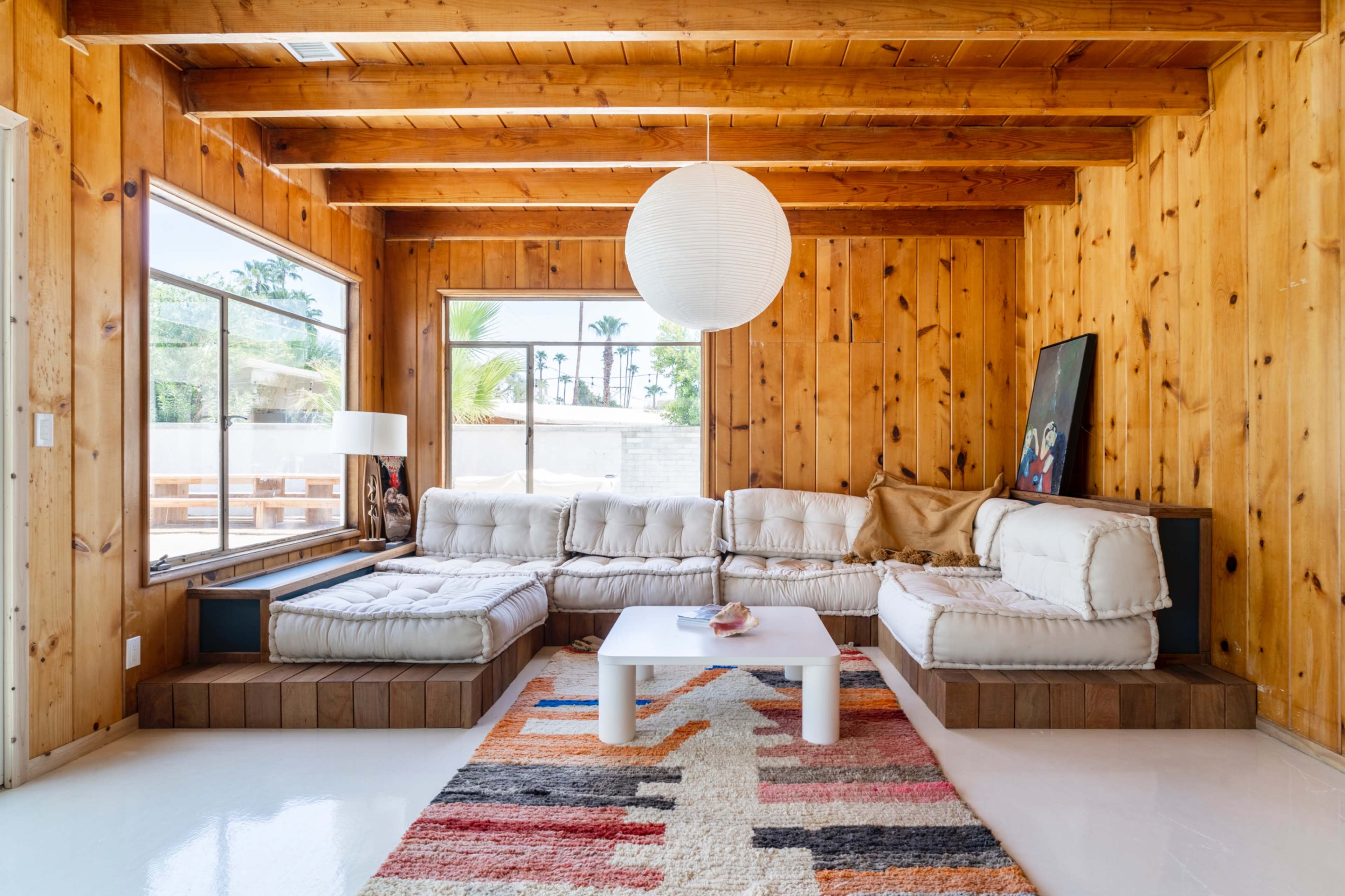 The scene features a living room with wooden paneling, a sectional sofa, a round paper lantern light fixture, and a colorful rug on a white floor.