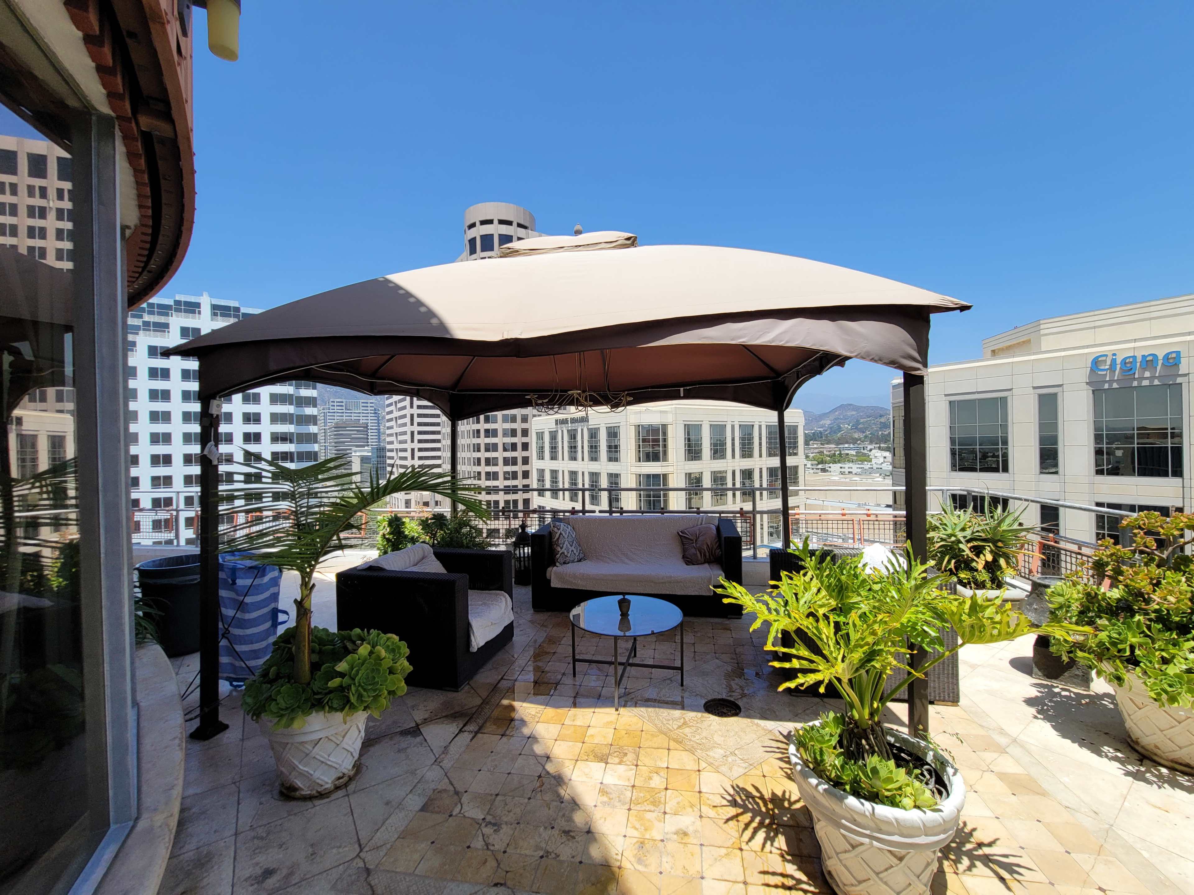 A shaded outdoor seating area with a gazebo, sofa, and plants overlooks a cityscape.