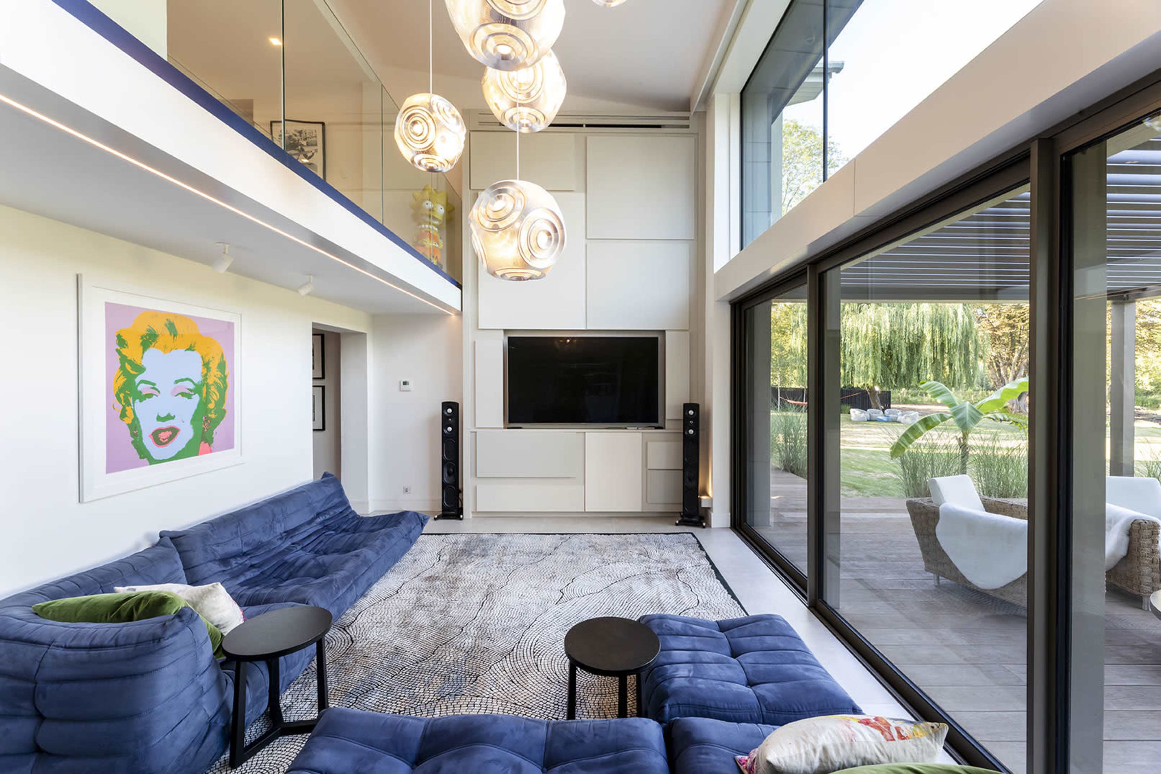 The image shows a modern living room with blue sectional sofas, a large flat-screen TV mounted on a white wall, and large glass windows providing a view of the outdoors.
