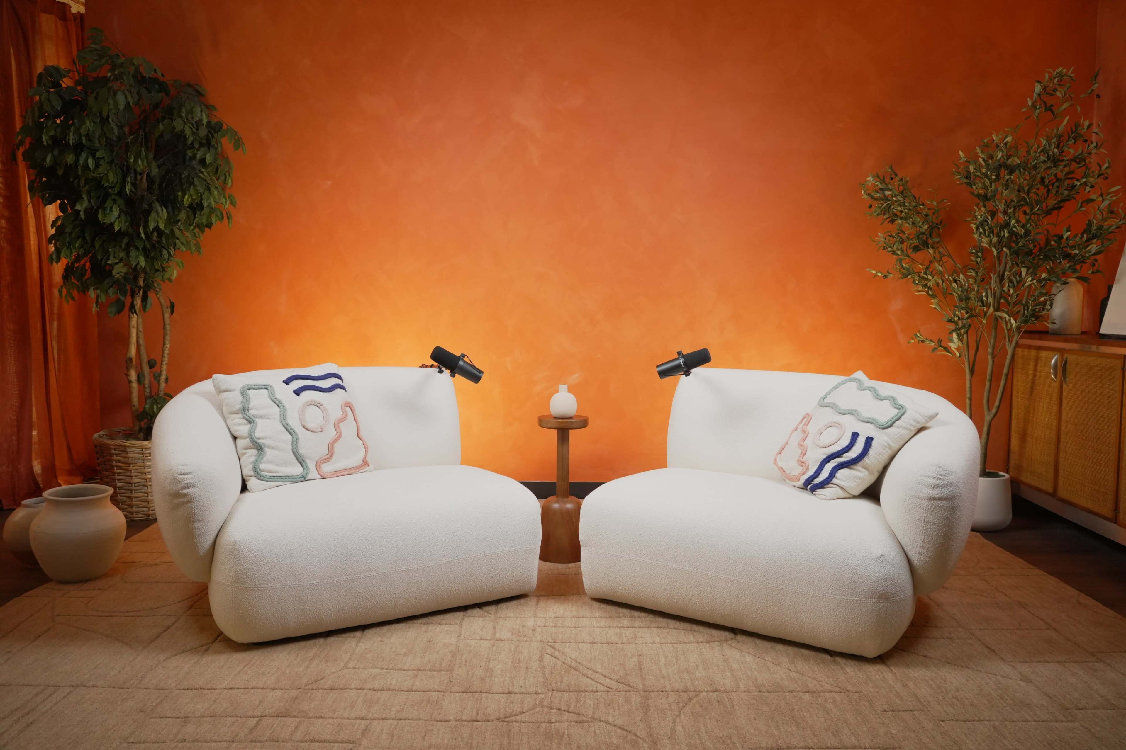 The image shows two white, plush chairs facing each other, with a small round table and decorative plants against an orange wall.